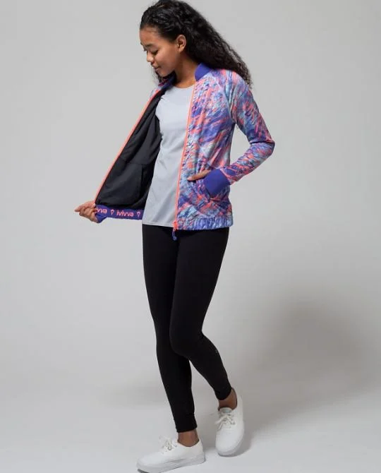 A young woman in a colorful patterned jacket, gray t-shirt, black pants, and white sneakers standing against a plain gray background, looking down and holding the edge of her jacket.
