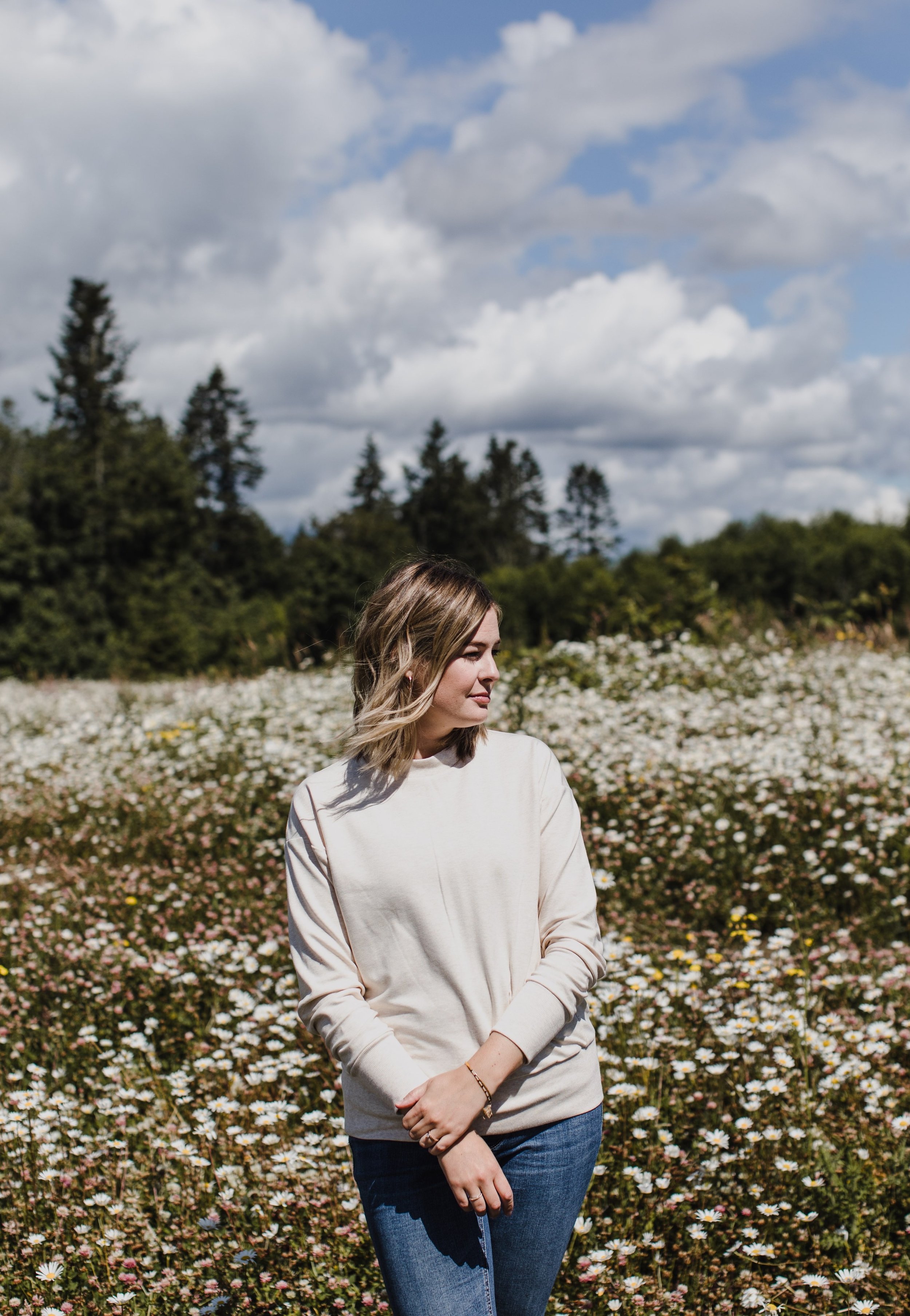 Young woman with light brown hair standing in a field of white daisies, trees and a partly cloudy sky in the background.