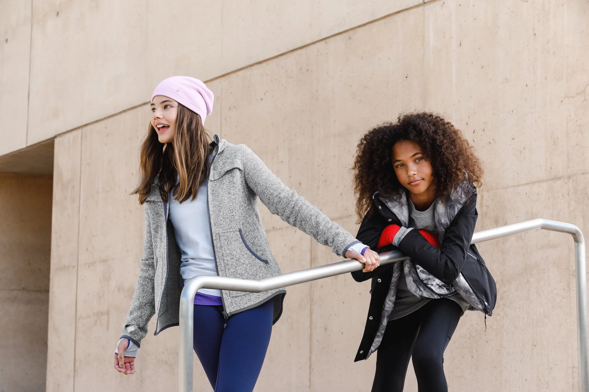 Two young women with curly and straight hair are leaning against a metal railing outdoors in front of a beige concrete wall. One woman is wearing a pink beanie and a grey jacket, smiling and looking to the side. The other woman is wearing a black jacket, resting her arm on the railing, and looking at the camera with a neutral expression.