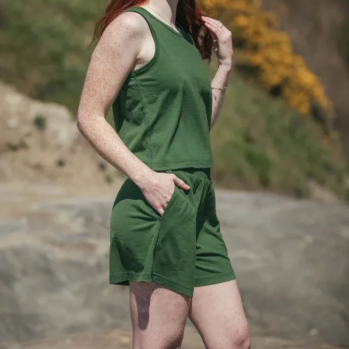A woman with freckles wearing a green tank top and matching green shorts, standing outdoors with a blurred natural background.