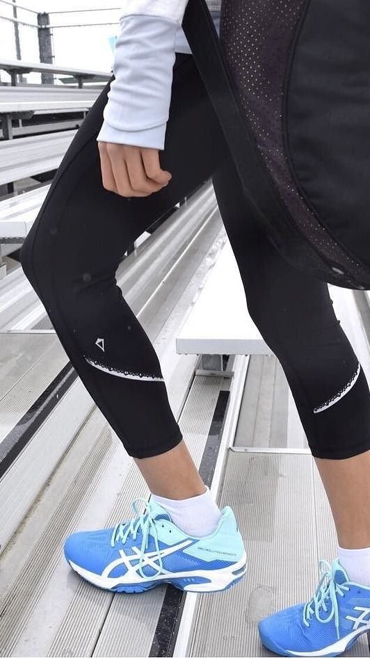 Close-up of a person wearing blue running shoes, black leggings with white accents, a white long-sleeve shirt, and a black jacket, standing on metal bleachers at an outdoor sports facility.