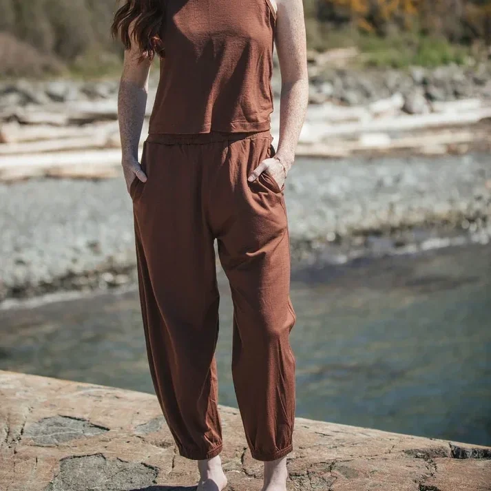 Person standing on rocks near a body of water, wearing a brown sleeveless top and matching loose pants with hands in pockets.