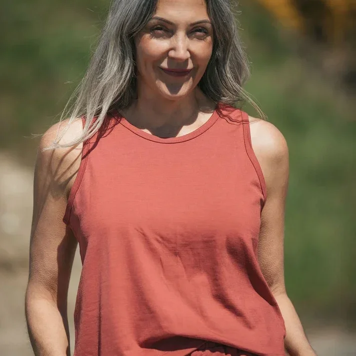 A woman with long, wavy gray hair wearing a sleeveless orange top outdoors with blurred green and autumn-colored trees in the background.