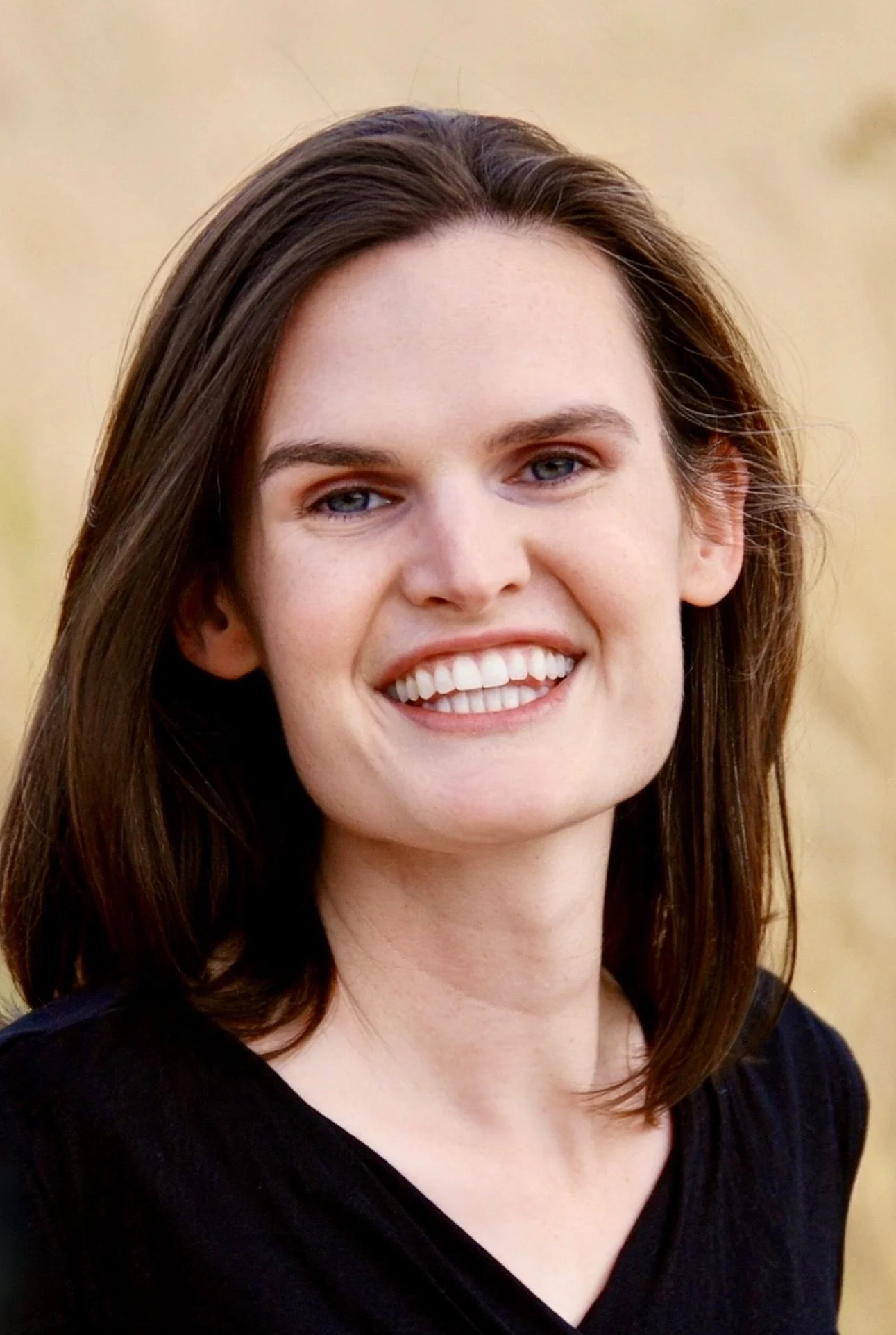 Close-up of a woman with shoulder-length brown hair, blue eyes, smiling, wearing a black top, with a blurred outdoor background.