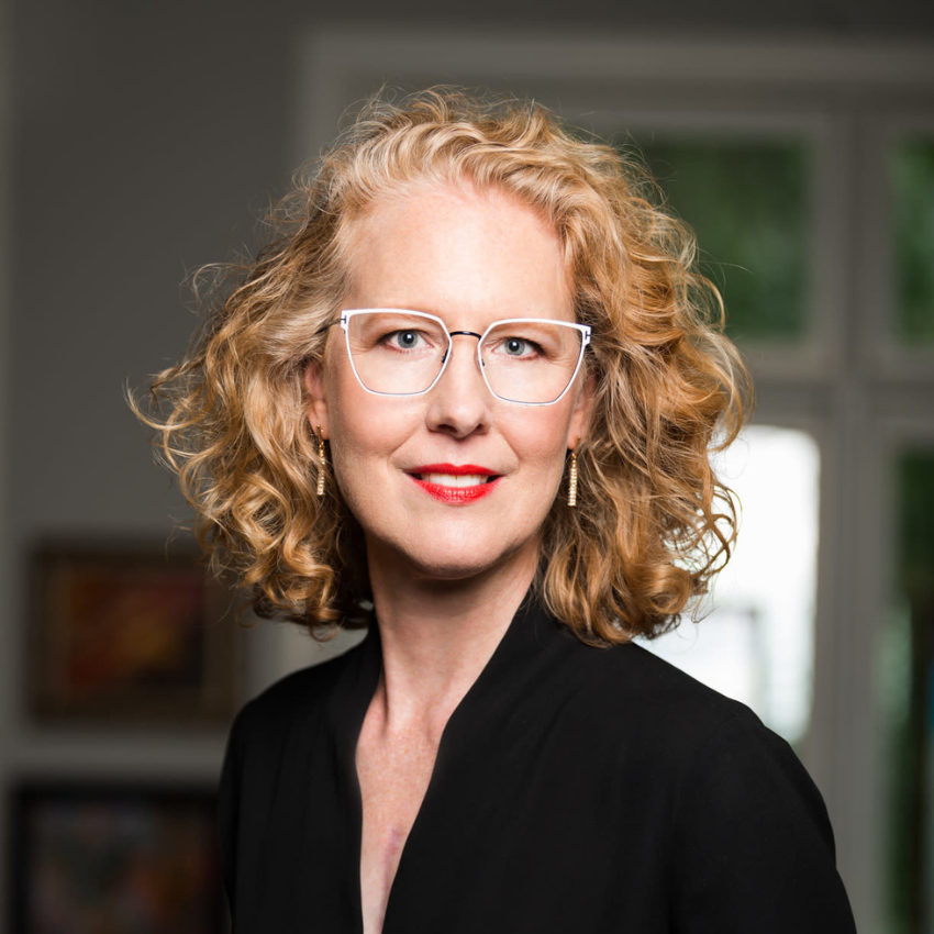 A woman with curly blonde hair, glasses, red lipstick, and earrings, wearing a black top, standing indoors in front of a window with a blurred background.