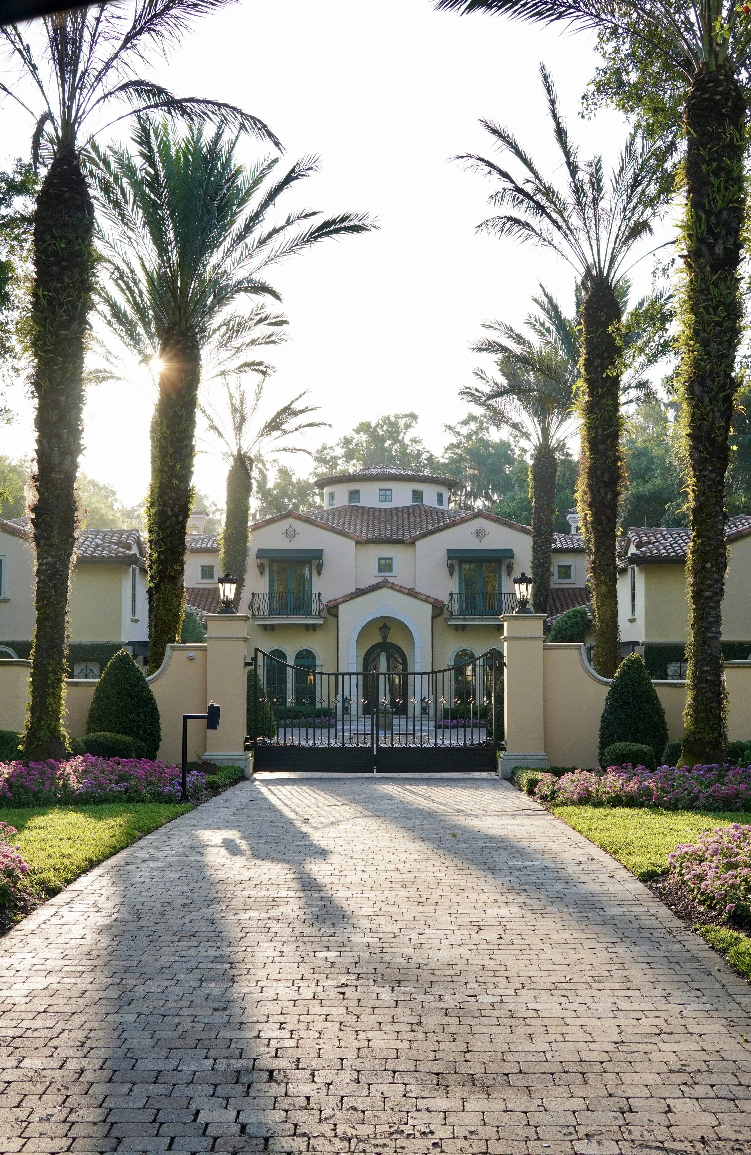 A white mansion with a tiled roof behind a tall black iron gate. Palm trees line the driveway, which is paved with light-colored bricks. Pink and purple flowers border the grassy areas on both sides of the driveway, and the sun is shining through the trees.