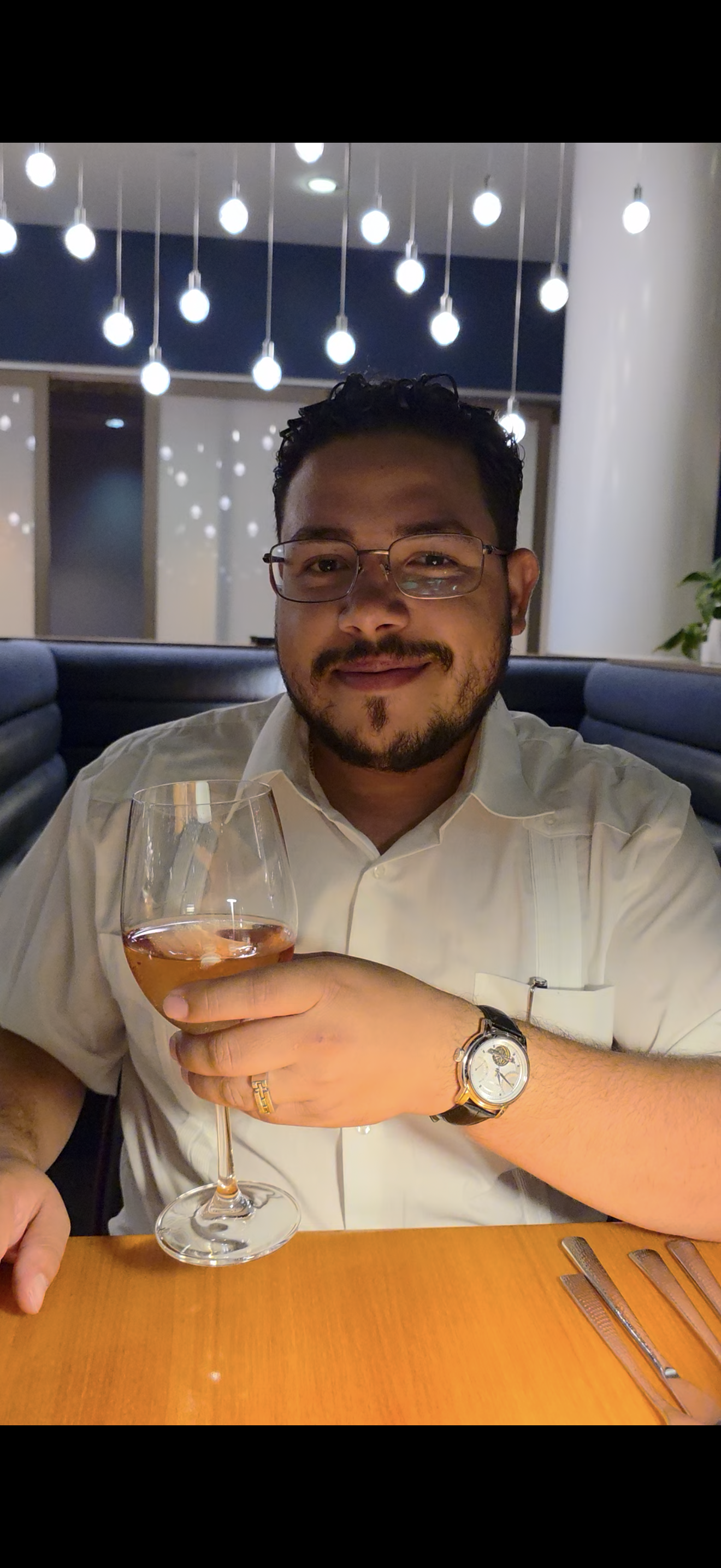 A man with glasses and a beard holding a glass of rosé wine in a restaurant. He is smiling, wearing a white shirt, a watch, and a ring. The background features modern overhead lighting with numerous hanging bulbs.