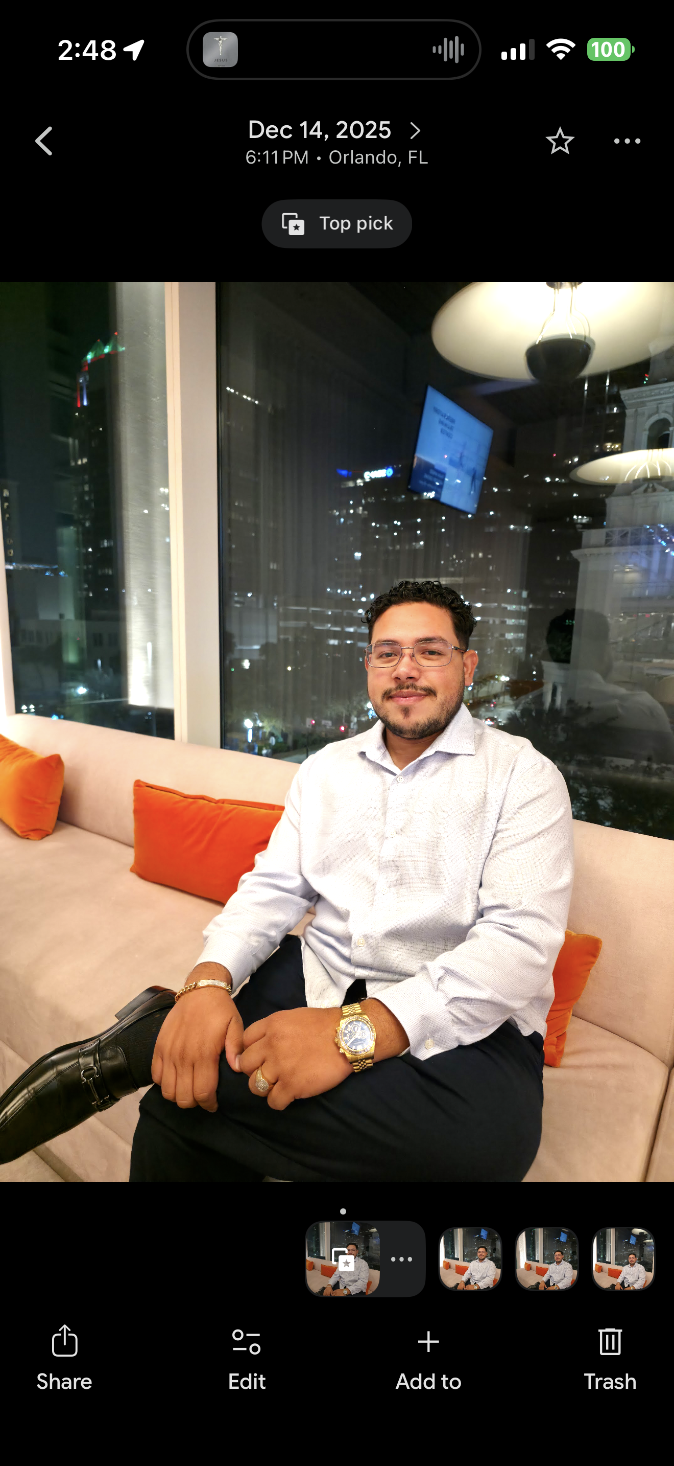 A man sitting on a beige couch with orange pillows in a modern, high-rise building at night. Behind him, large windows reveal a cityscape of tall buildings with illuminated windows. The man is dressed in a white shirt, black pants, and wearing glasses, a gold watch, and a ring. There are pendant lights and a TV mounted on the window reflection.