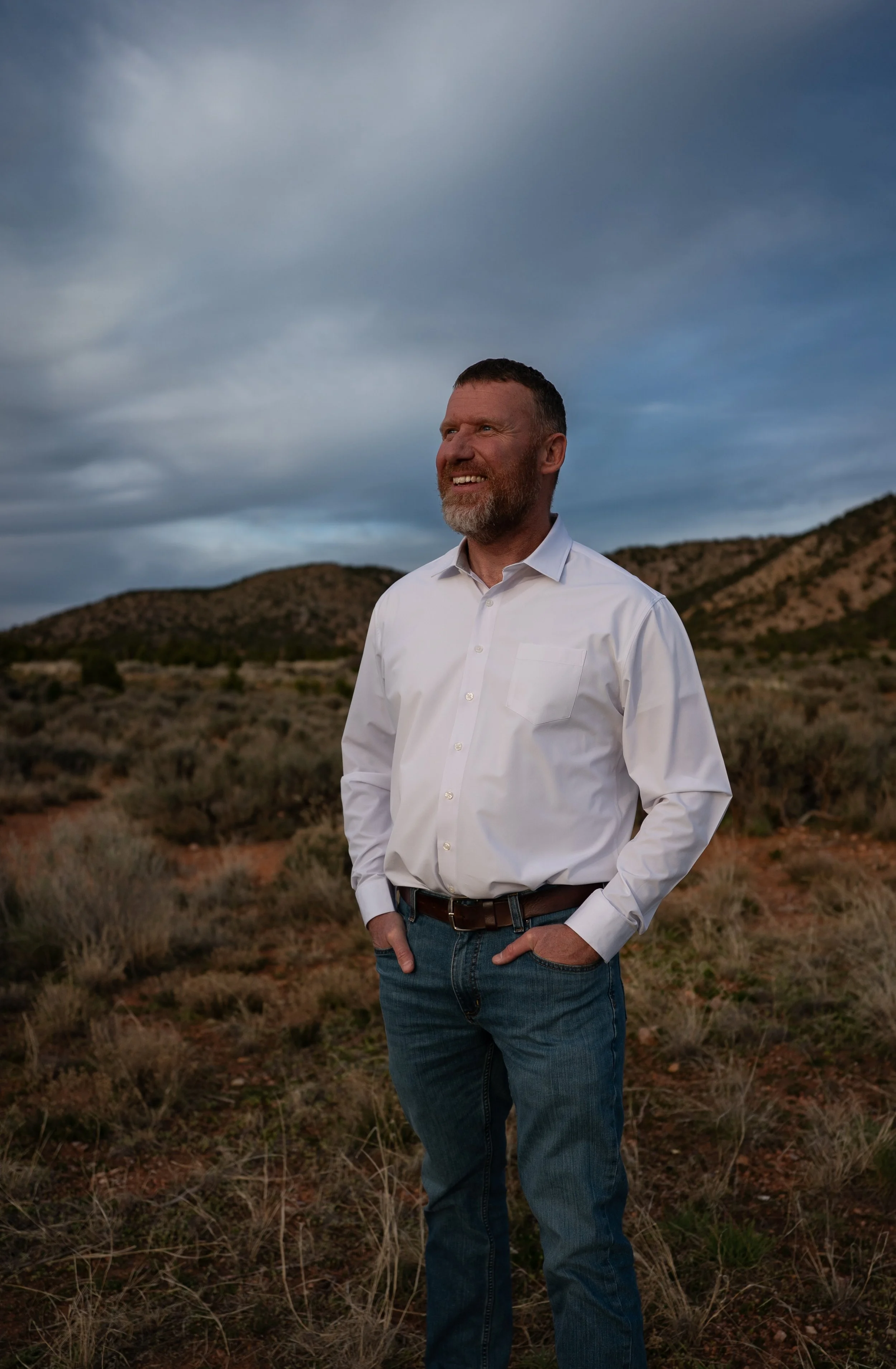 A man in a white shirt and blue jeans standing outdoors in a dry, shrub-covered landscape with mountains and cloudy sky in the background, smiling and looking to the side.