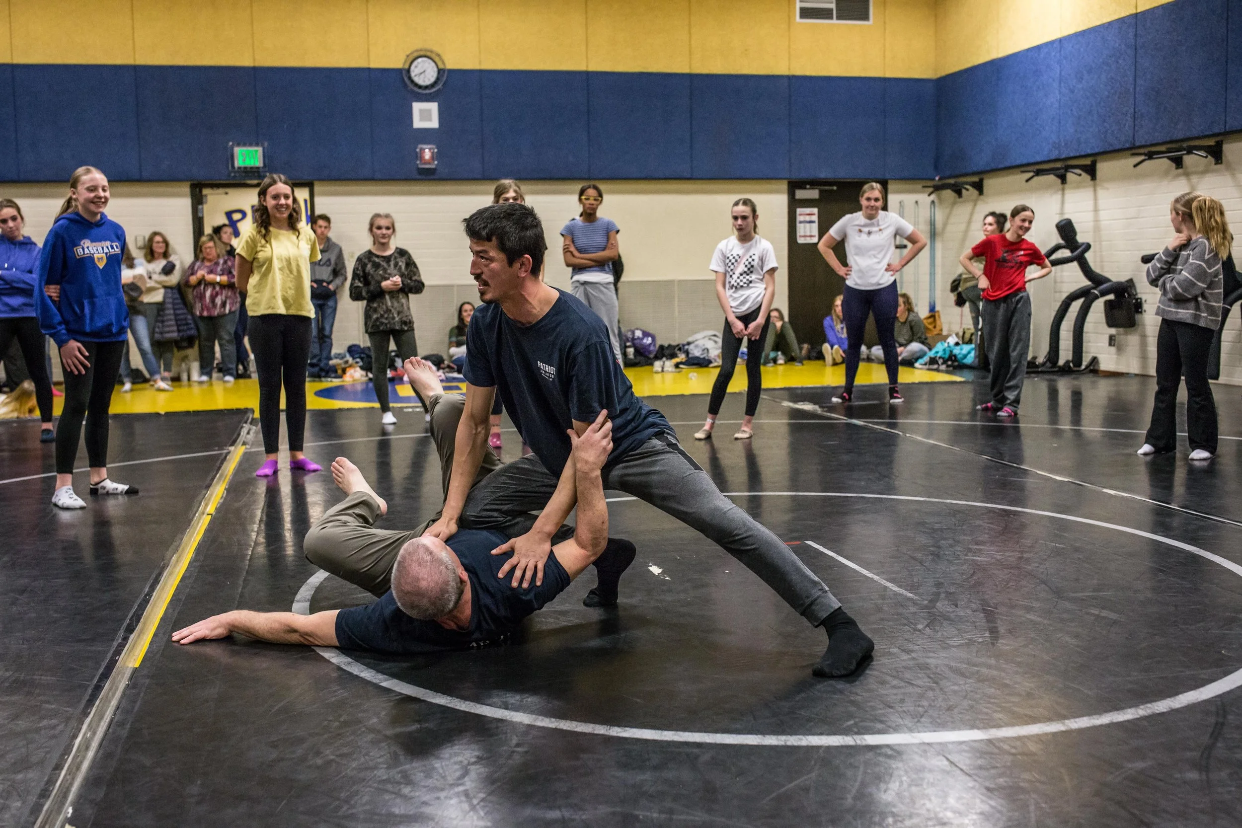A wrestling demonstration with one person on the ground in a grapple while another person is standing over him, surrounded by a group of onlookers in a gymnasium.