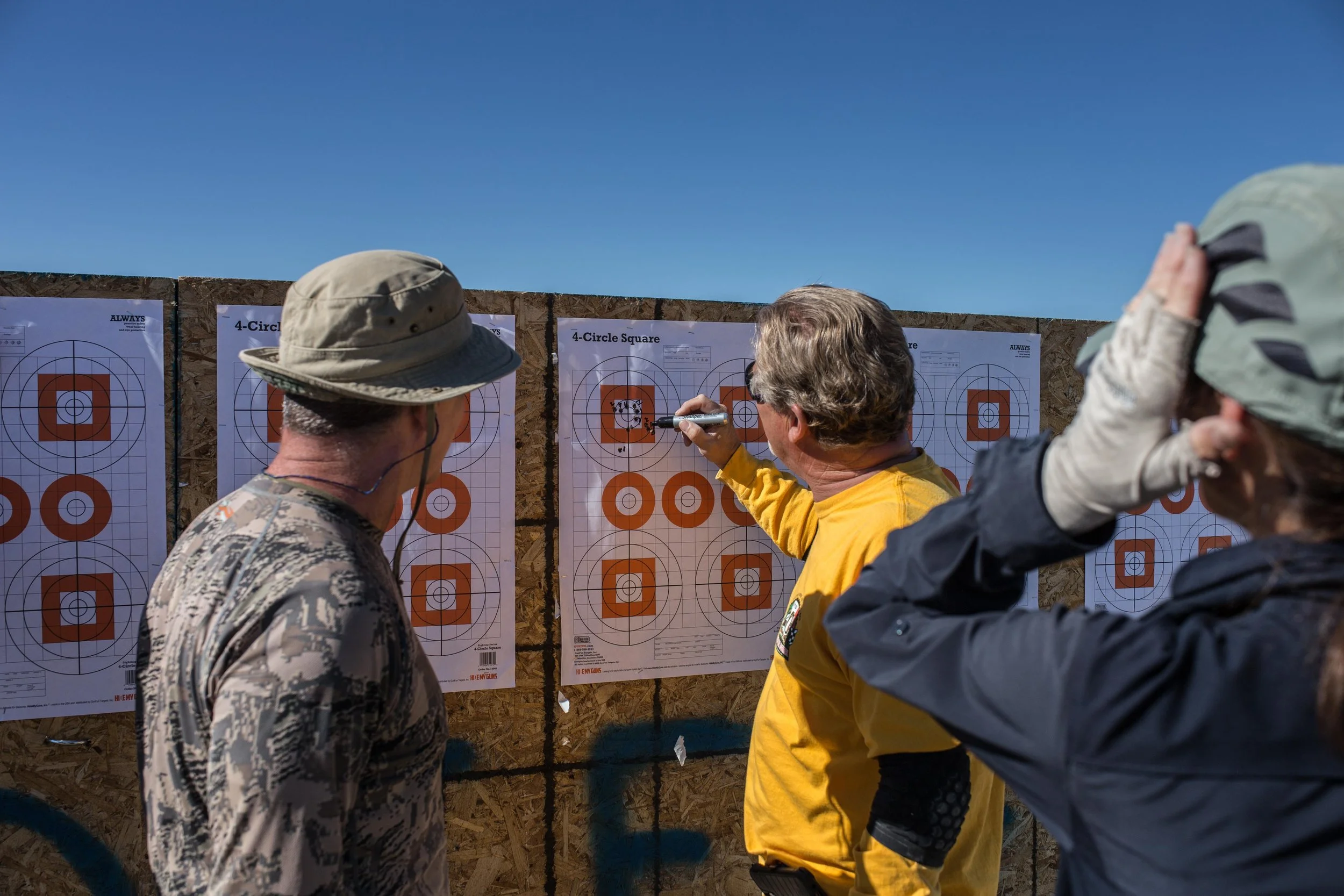 Three people at an outdoor shooting range examining target sheets with geometric shapes.