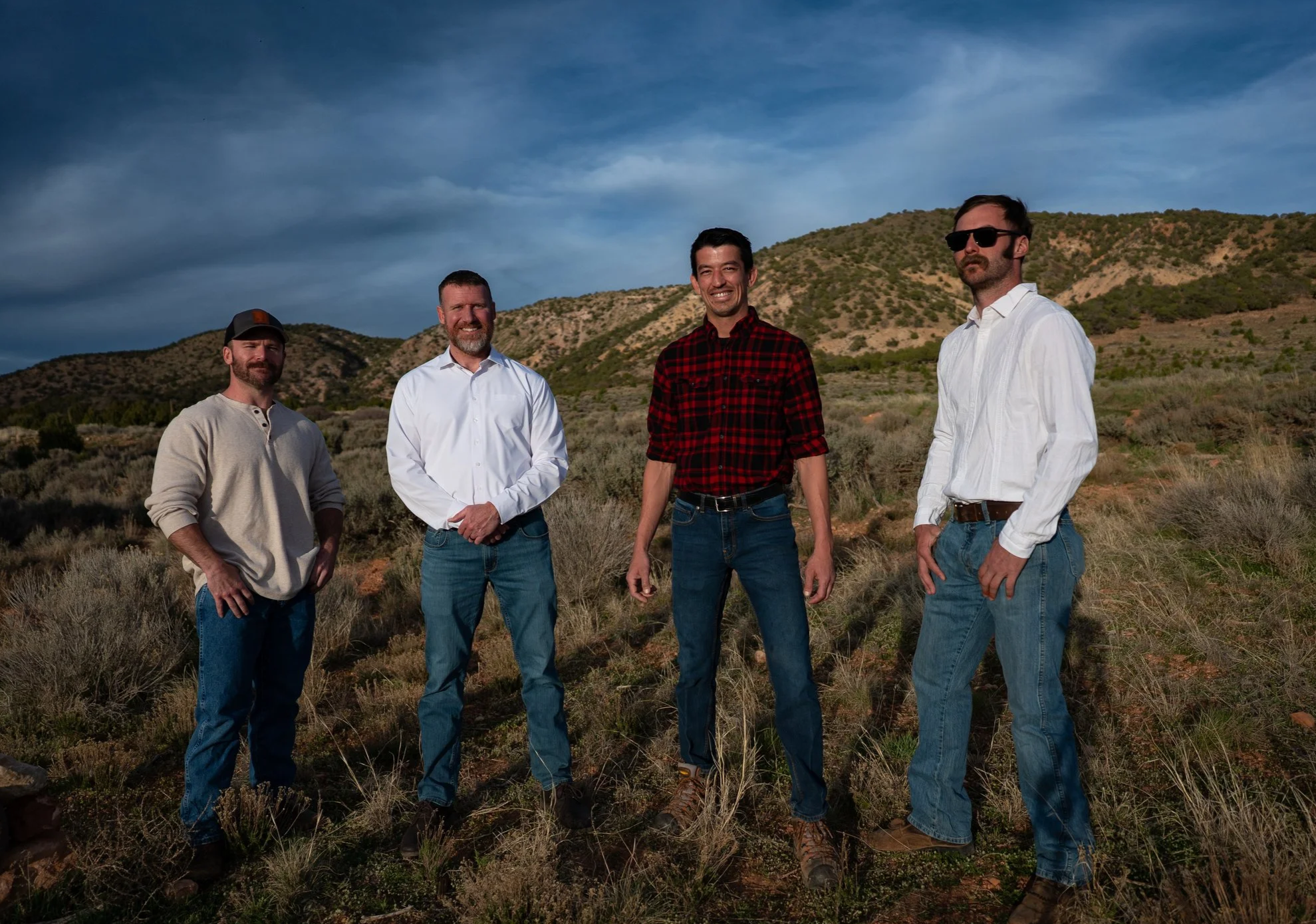 Four men standing outdoors in a grassy, hilly landscape under a cloudy sky, posing for a photo.