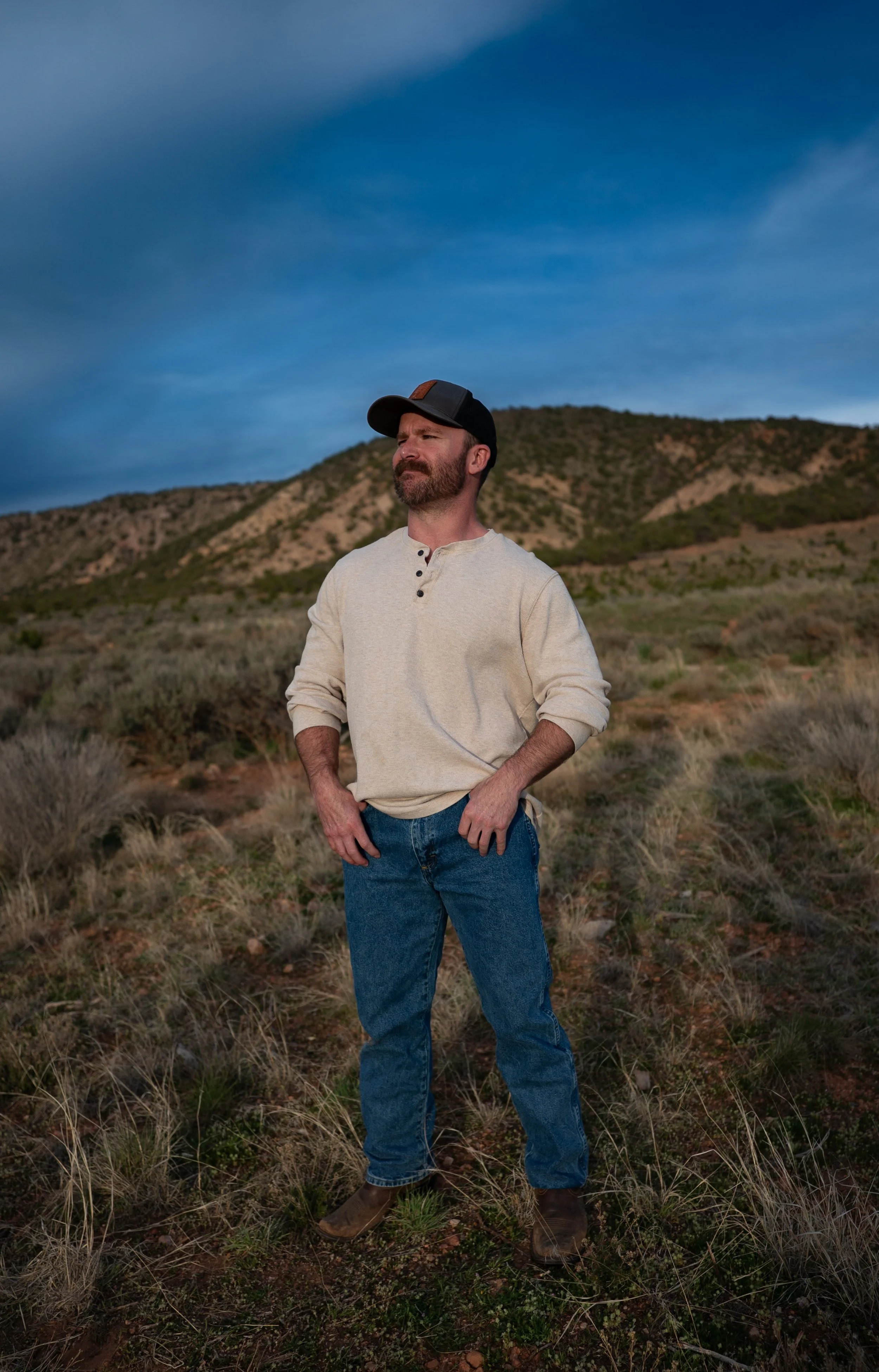 A man stands outdoors in a desert landscape with hills and sparse vegetation under a cloudy sky, wearing a beige long-sleeve shirt, blue jeans, brown boots, and a black baseball cap.