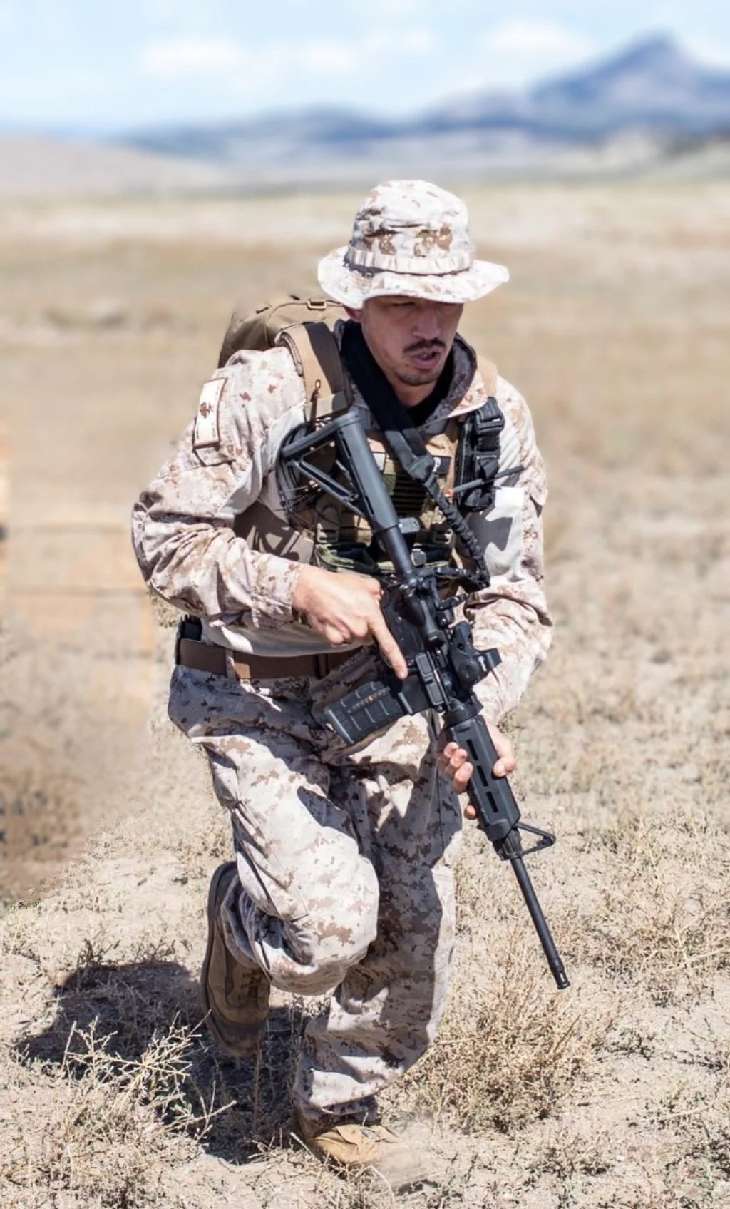 A soldier in desert camouflage uniform running in a dry, open field with mountains in the background, carrying a rifle and wearing a backpack.