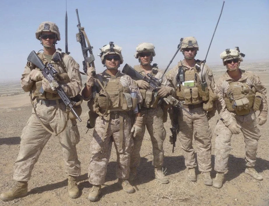 Six soldiers in desert camouflage uniforms and helmets standing together in a desert landscape, holding rifles and equipment.