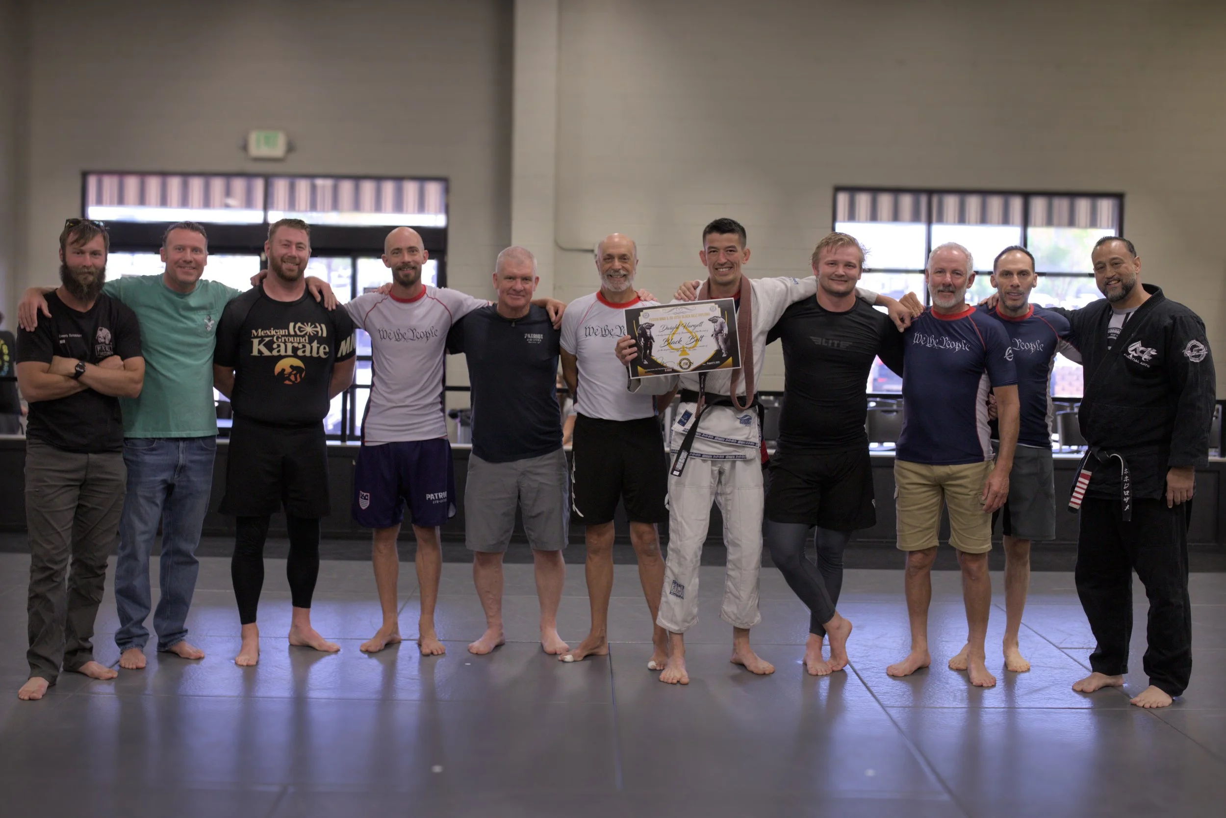 Group of eleven men standing in a martial arts training facility, with some wearing martial arts gis and others in casual athletic clothing. They are smiling, with their arms around each other's shoulders, and one is holding a framed certificate or award.