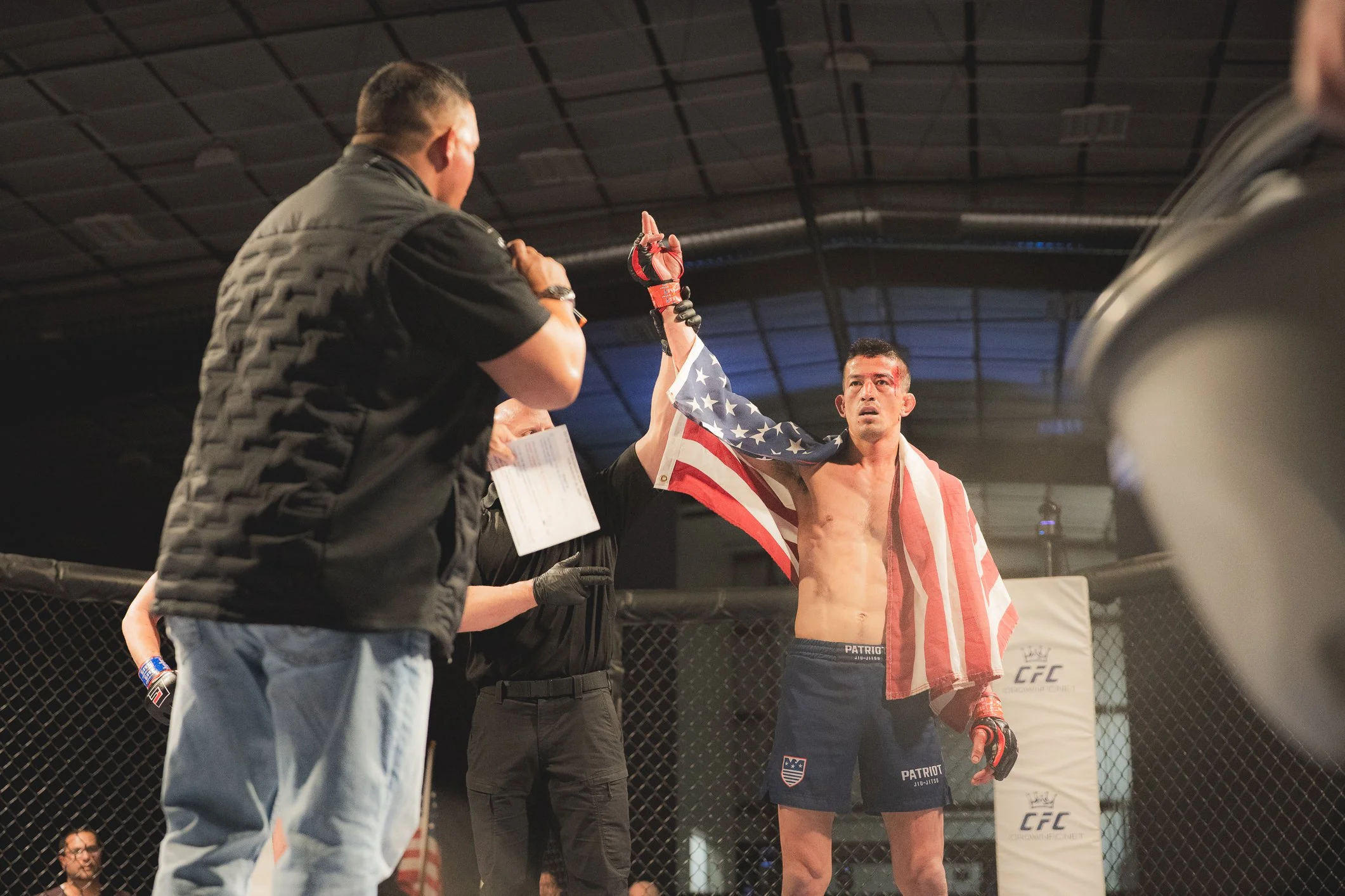 Mixed martial arts fighter with an American flag wrapped around his shoulders, raising his hand in victory inside the cage, with officials and a referee present.