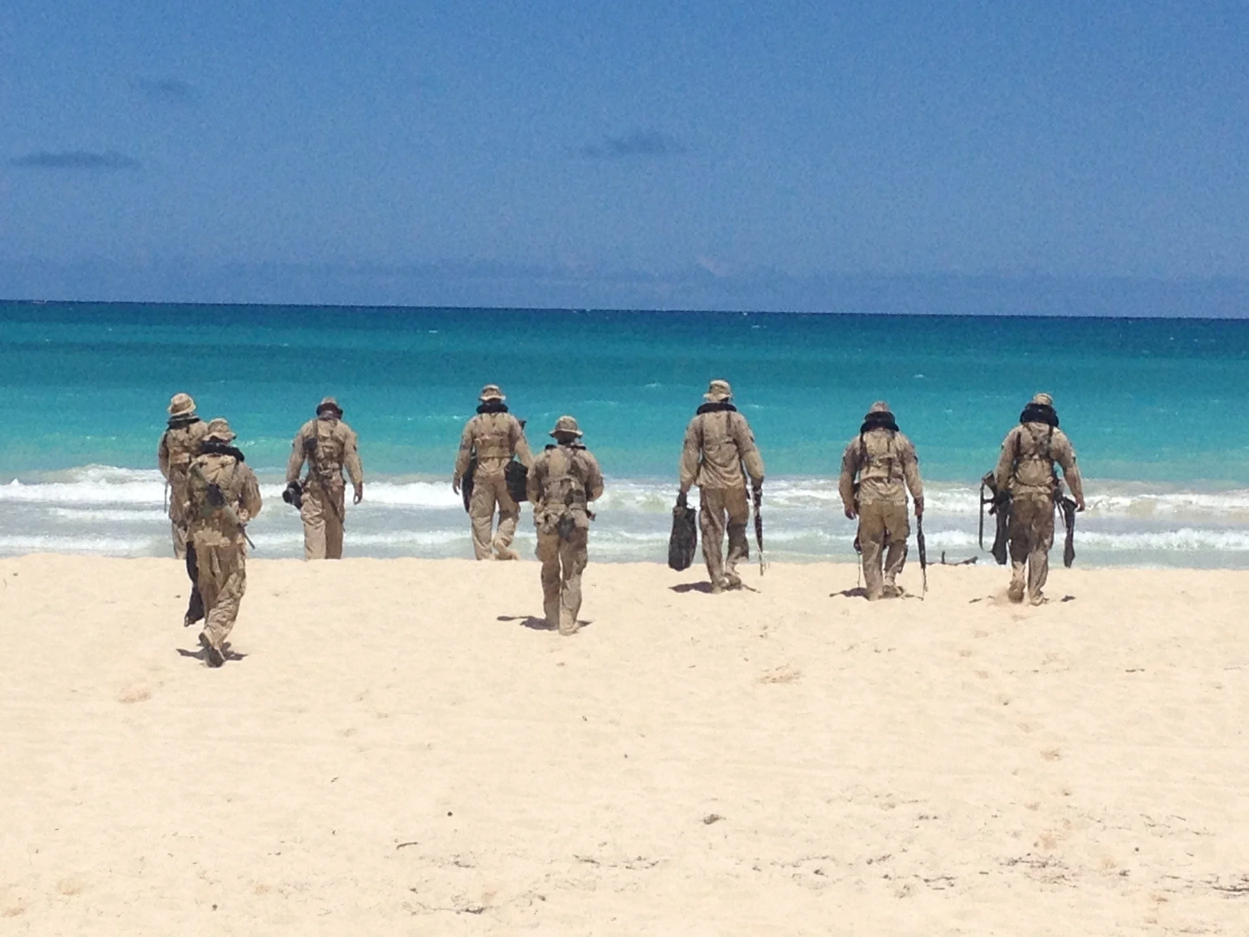 A group of nine soldiers in desert camouflage uniforms walking towards the ocean on a sandy beach, carrying equipment and gear under a blue sky.