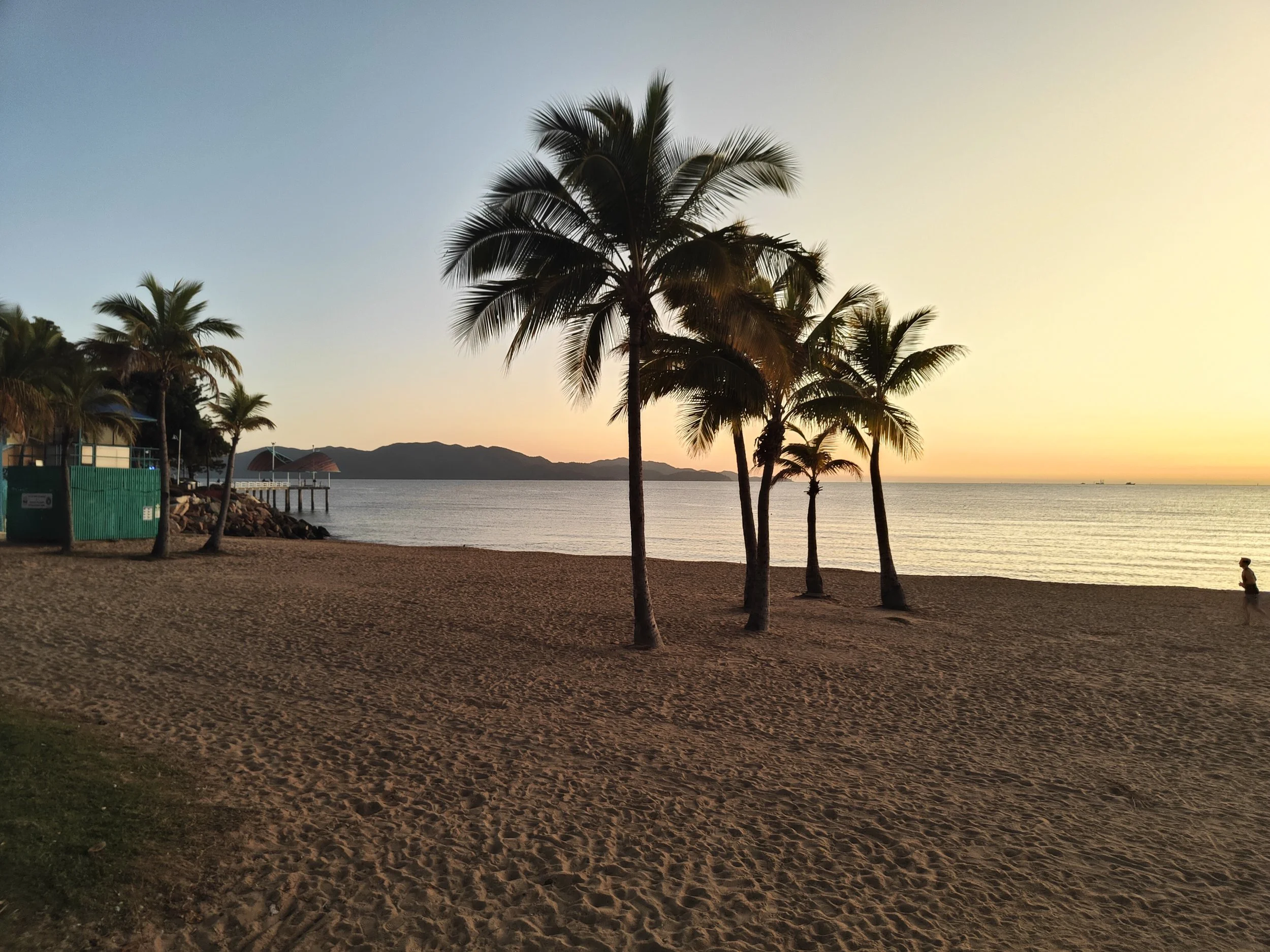 A beach scene during sunset with palm trees, calm water, a pier, and mountains in the distance, with a person standing on the sand.