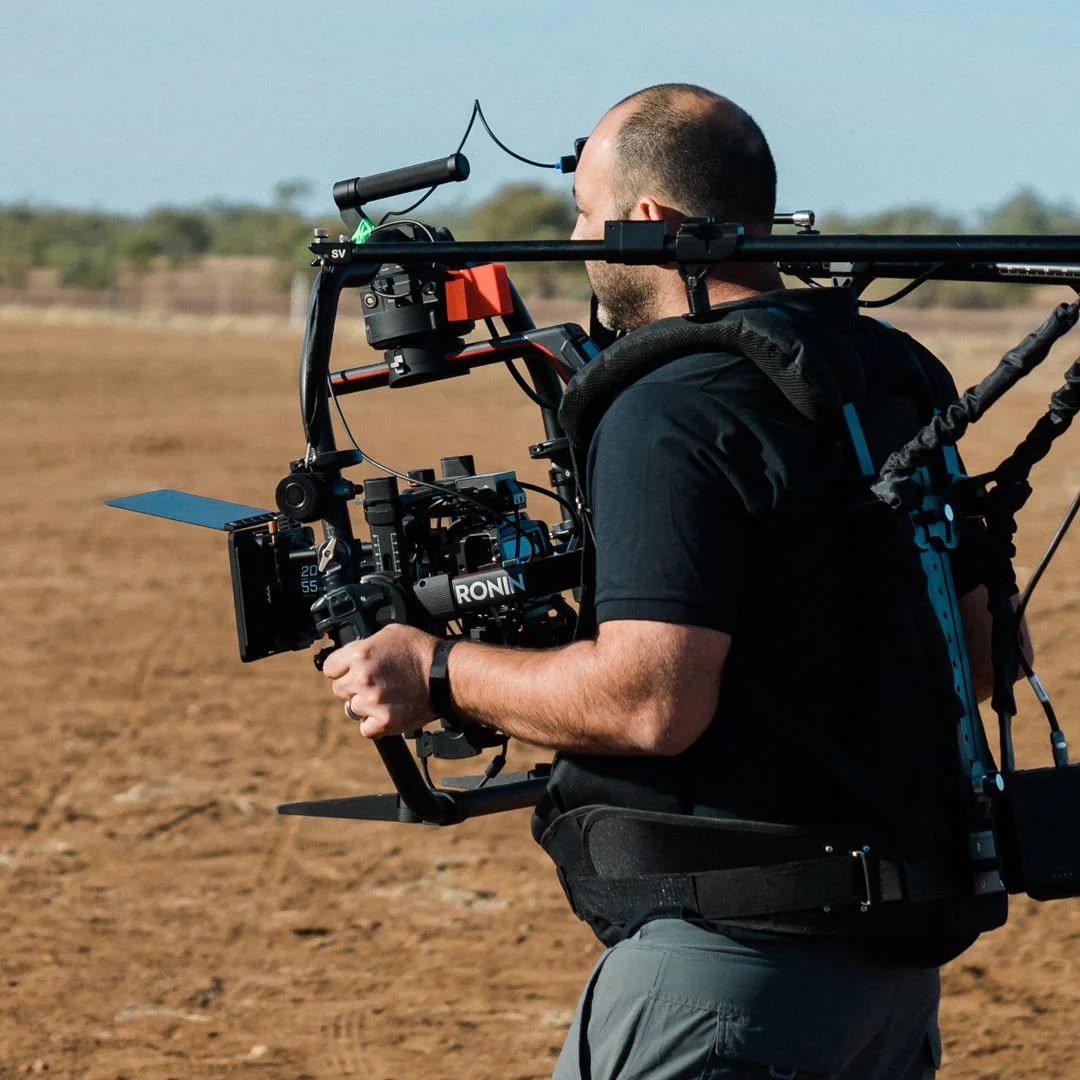 A man operating a professional camera mounted on a stabilizer rig outdoors in a desert landscape.