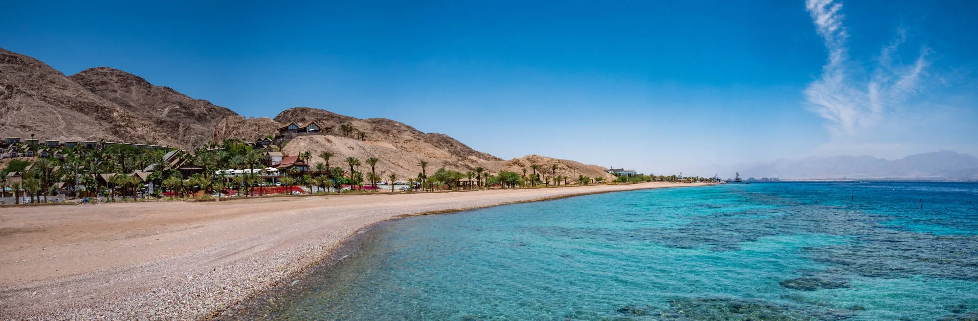 View of a coastal beach with turquoise water, sandy shore, palm trees, and buildings against arid mountains under a clear blue sky.