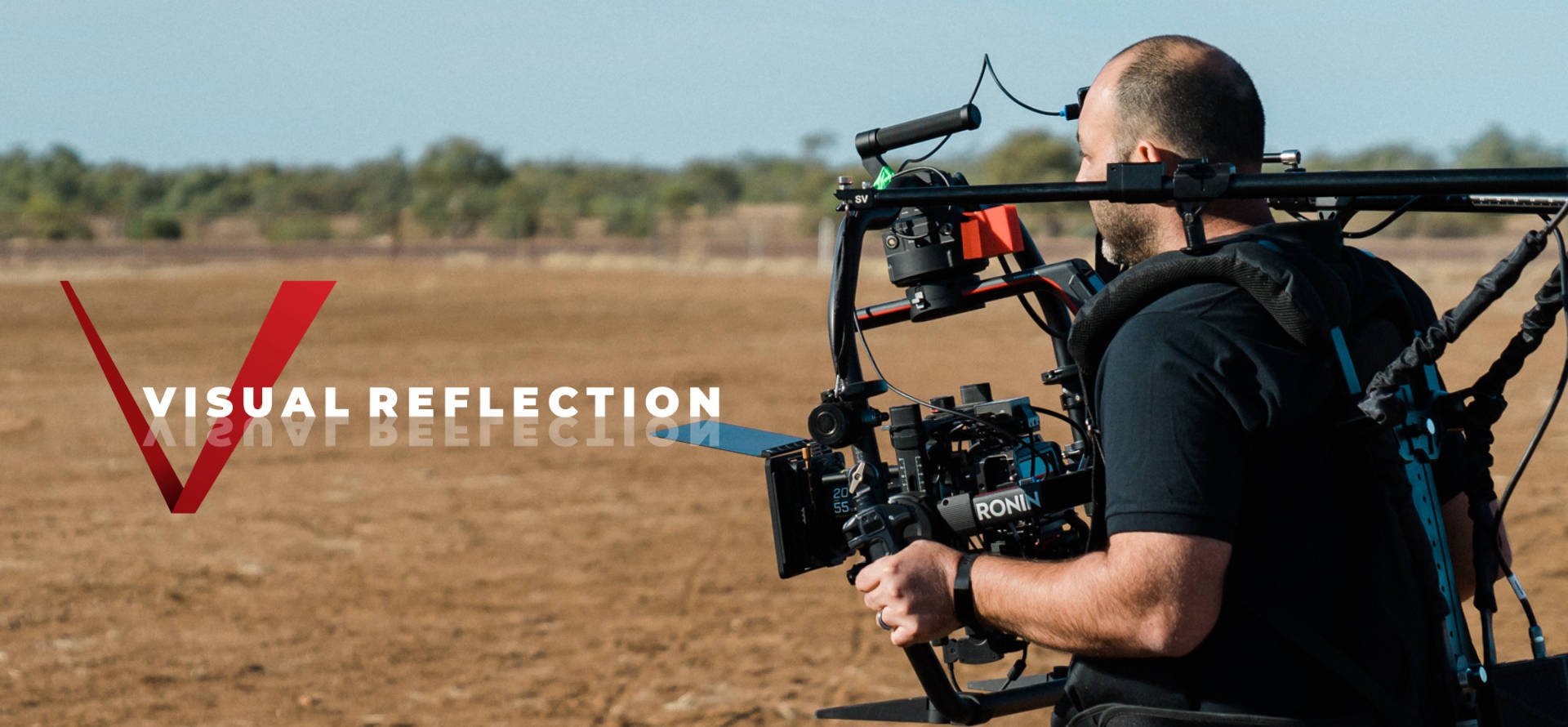 Man operating a camera mounted on a steady cam rig outdoors in a field with sparse trees in the background, with the words "V Visual Reflection" in the lower left corner.