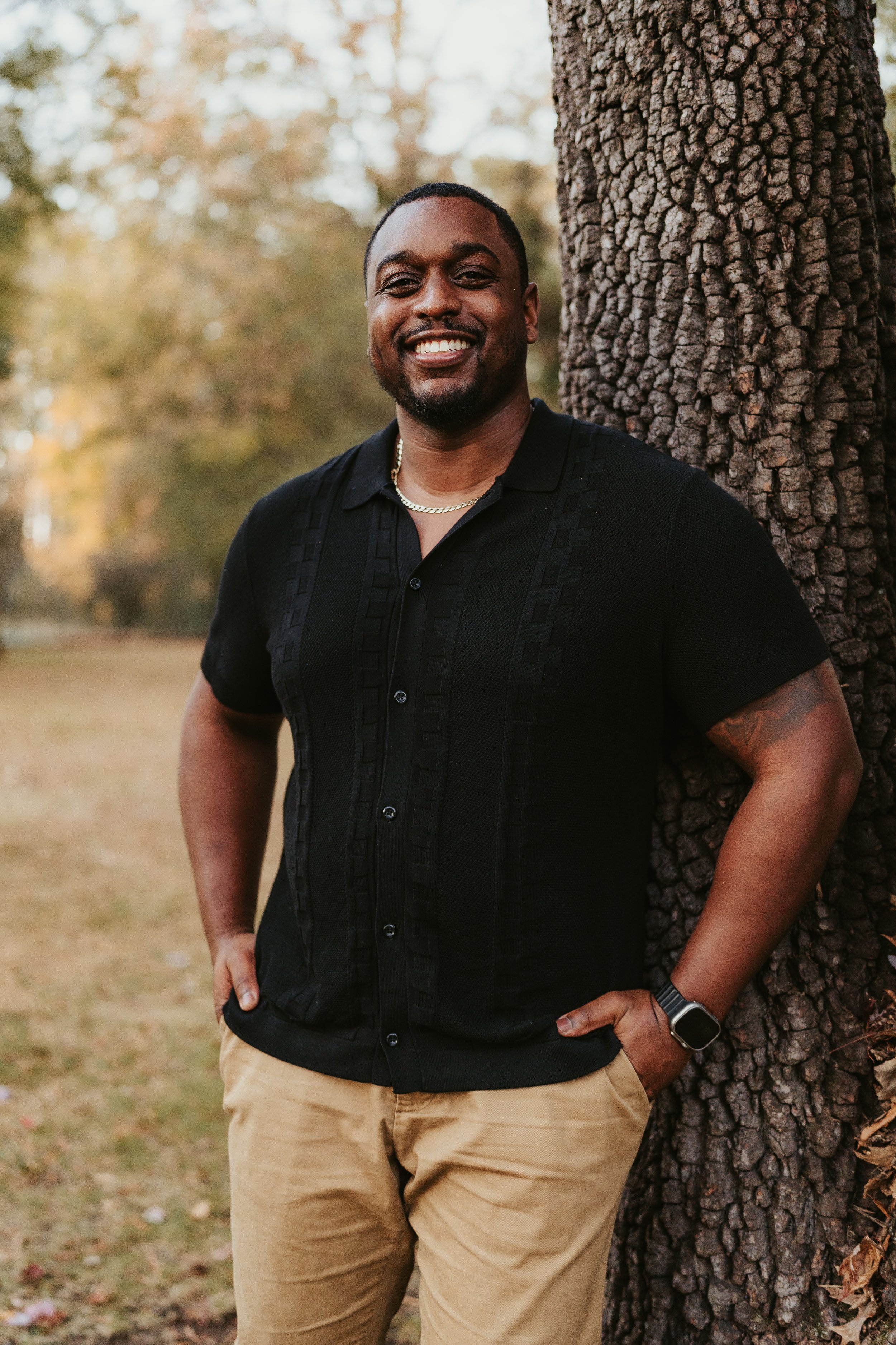 A smiling man is standing outdoors next to a tree, wearing a black short-sleeved button-up shirt, beige pants, a gold necklace, and a watch.