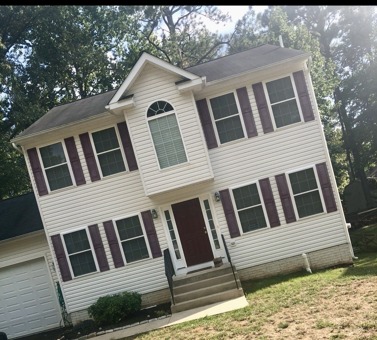 Front view of a two-story house with white siding, red shutters, and a red front door, surrounded by green trees and lawn. After being soft washed