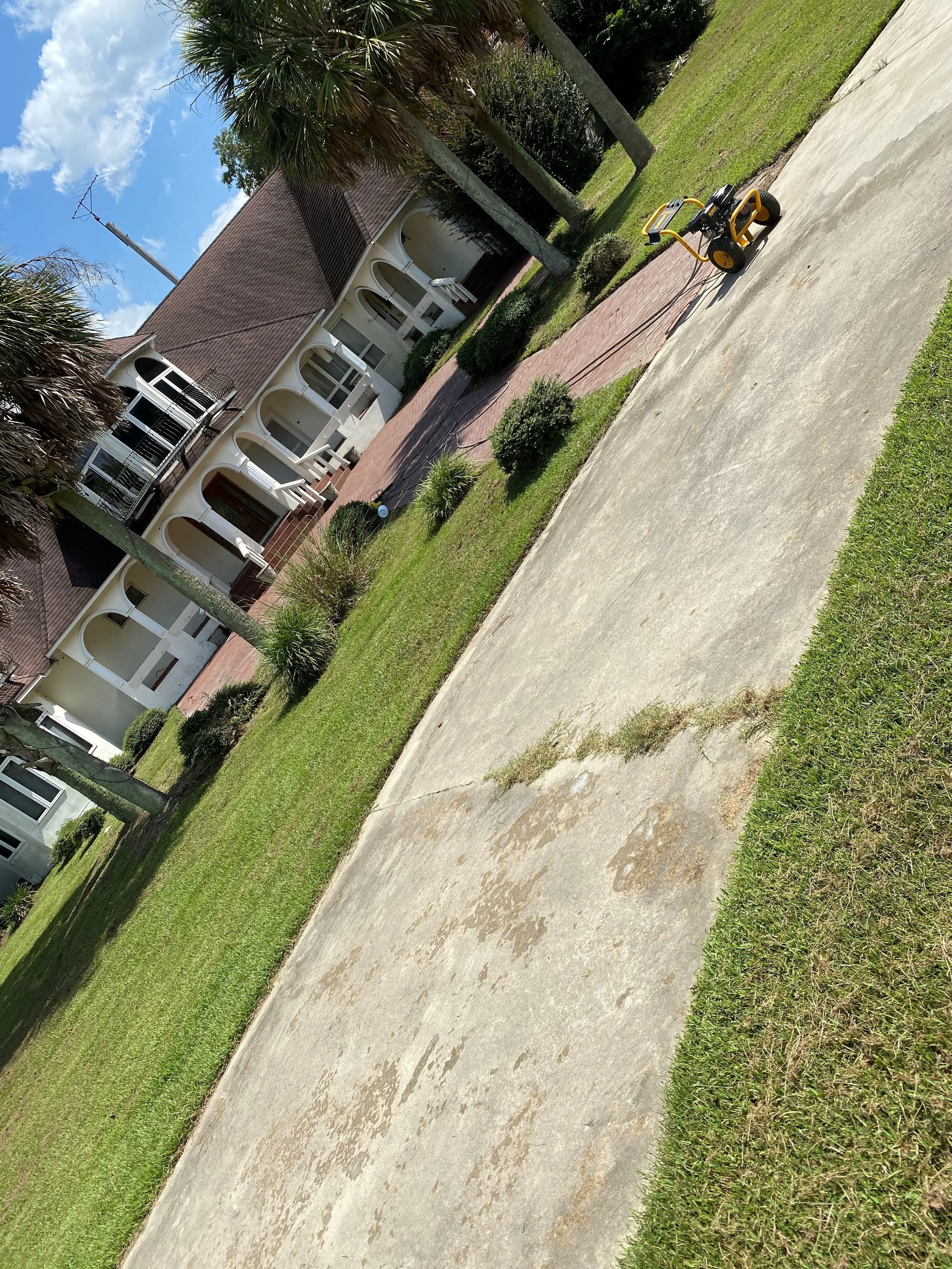 A large concrete driveway, a yellow pressure washer, and a well-maintained lawn with small bushes. In the background, there is a white house with balconies and arches. Before being pressure washed