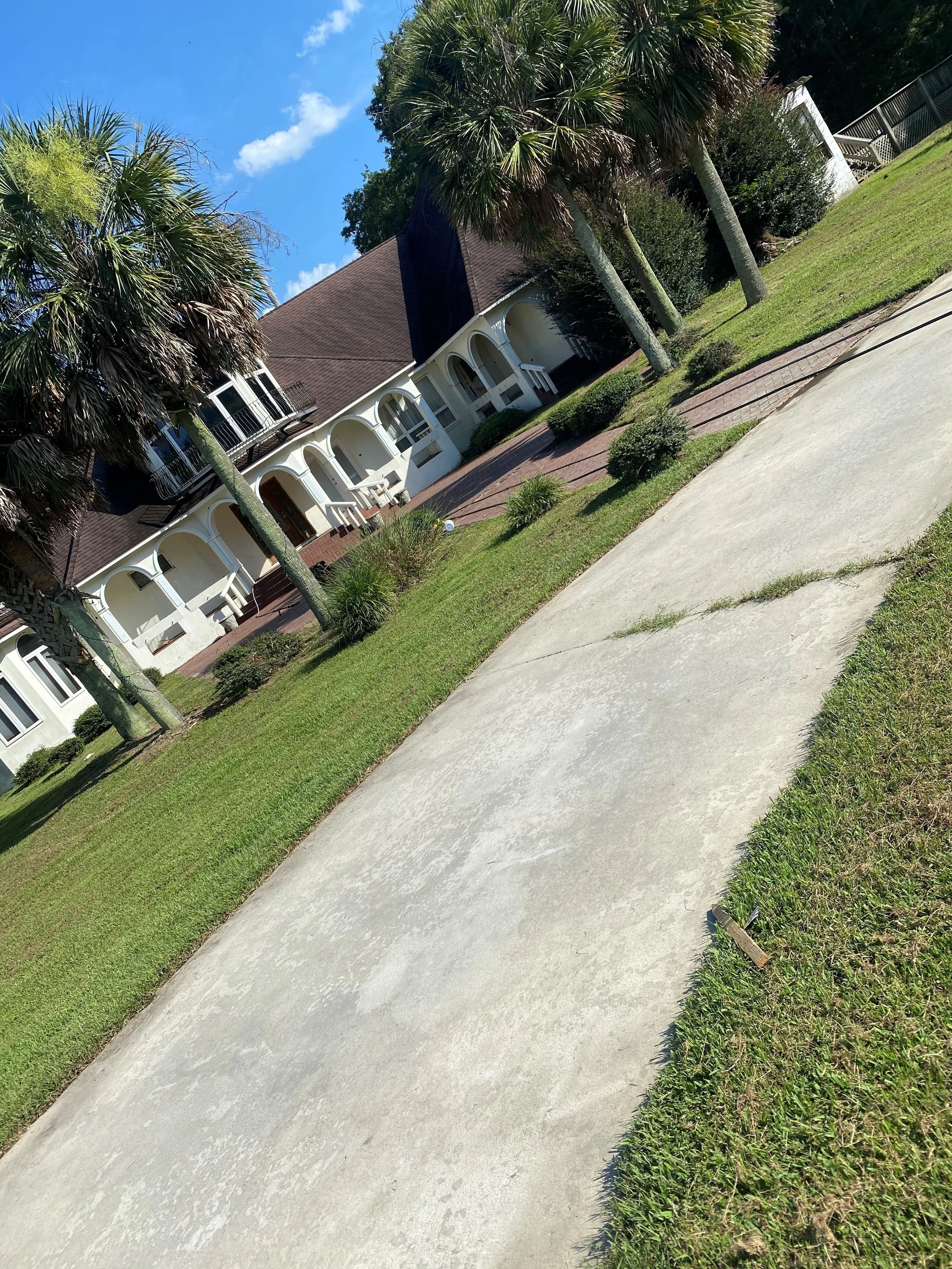 A large concrete driveway in front of a large white house with a brown roof, arched windows, and balconies, surrounded by palm trees. After being pressure washed