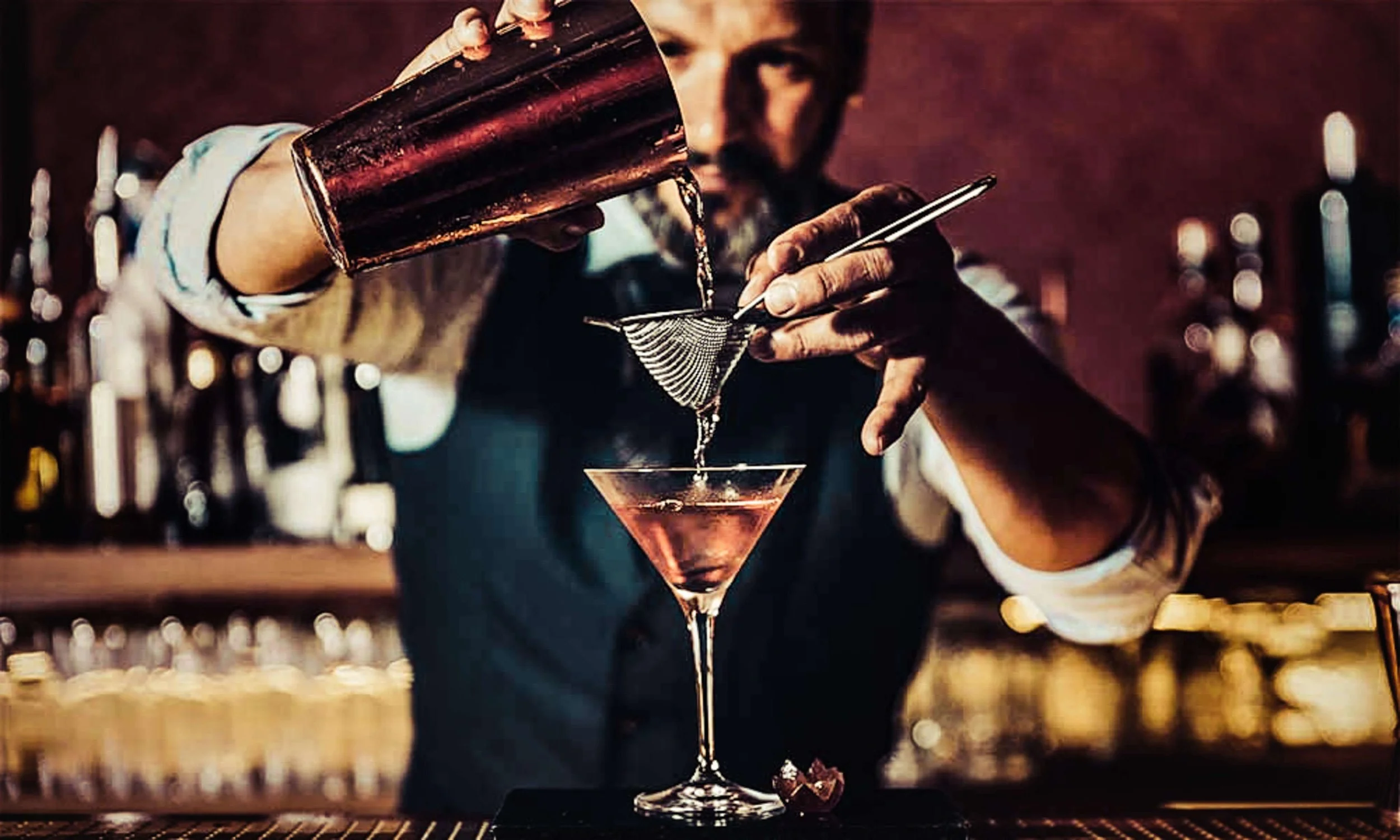 A professional bartender in a vest straining a cocktail from a copper shaker into a martini glass.