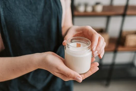 A close-up shot of a person wearing a dark apron and holding a handcrafted white soy candle in a clear glass jar with a wooden wick.