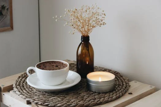 A cozy still life on a wooden surface featuring a white cup of coffee with foam, a lit candle in a silver tin, and a small amber glass bottle holding dried white flowers on a woven jute placemat.