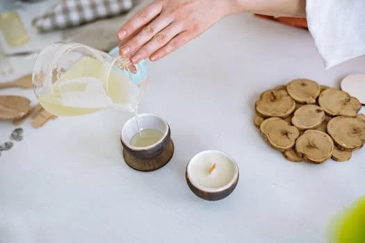 A person's hand pours melted pale yellow wax from a glass pitcher into a small, dark ceramic candle vessel. Another finished white candle with a wooden wick sits nearby on a white workspace next to a pile of small wood slices.