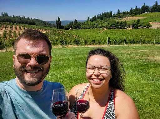 A man and woman smiling outdoors, holding glasses of red wine, with a vineyard and green hills in the background.