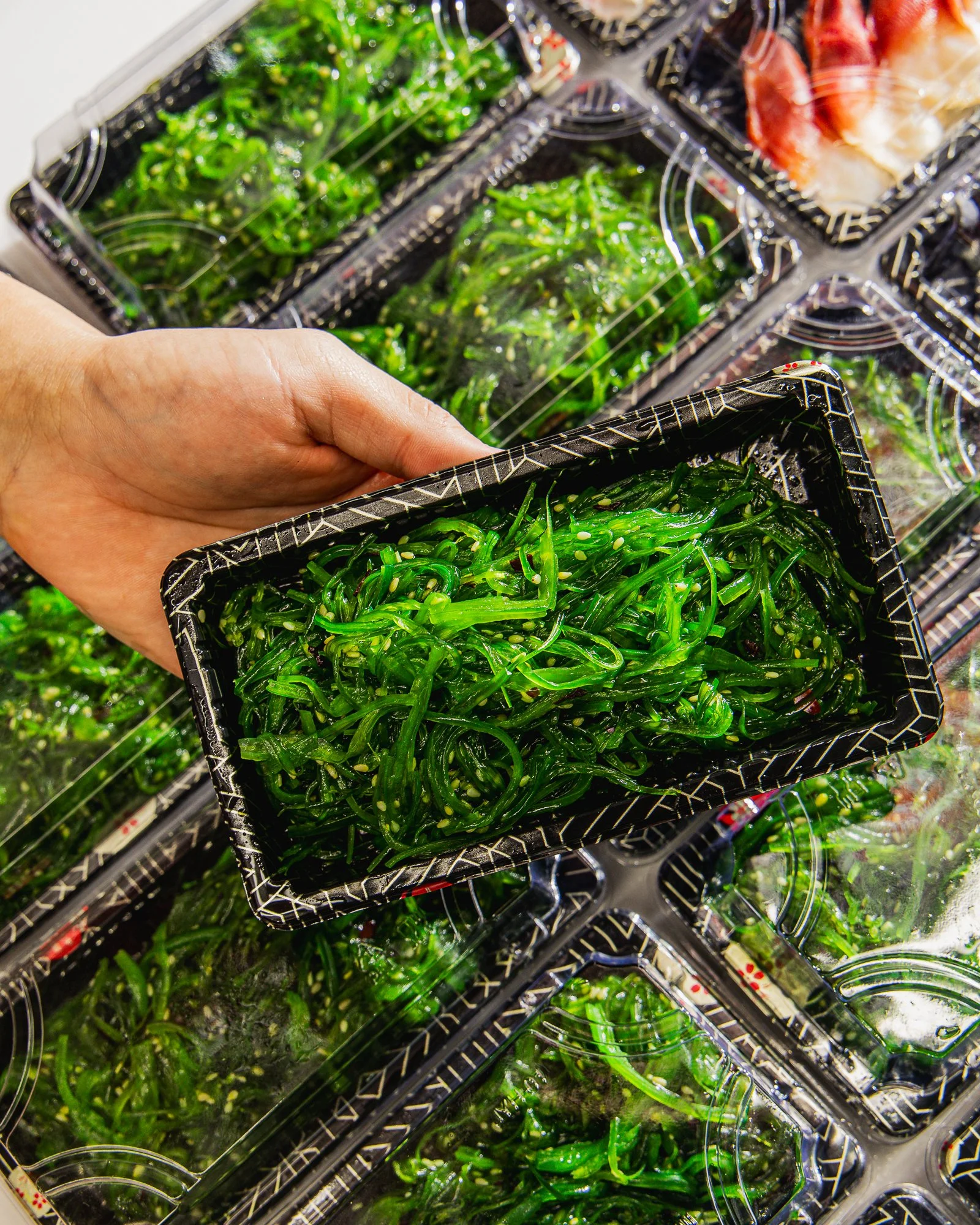 Close-up of a hand holding a black plastic container filled with green seaweed salad, with more containers of seaweed salad stacked behind.