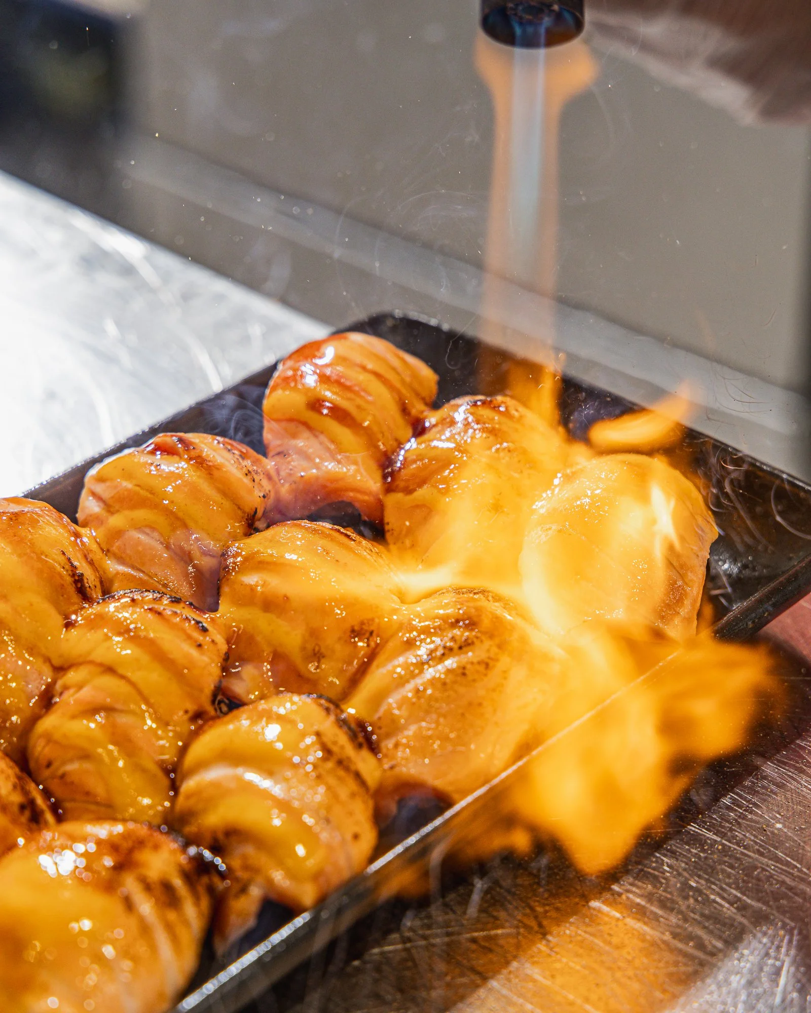 Close-up of skewered grilled meatballs with melted cheese, on a black tray over a wooden surface.