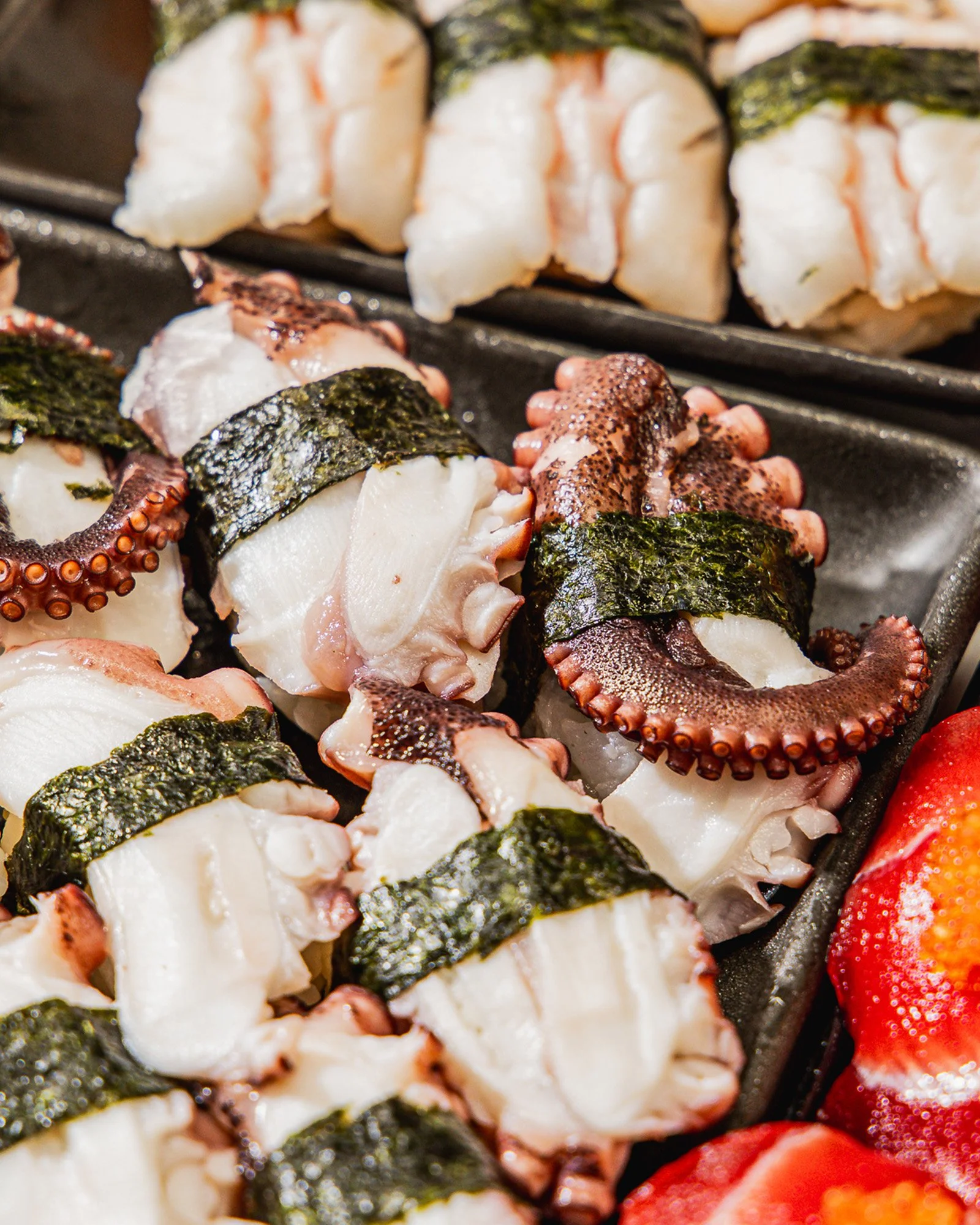 Close-up of sushi rolls with octopus, seaweed, white rice, and strawberries on a tray.