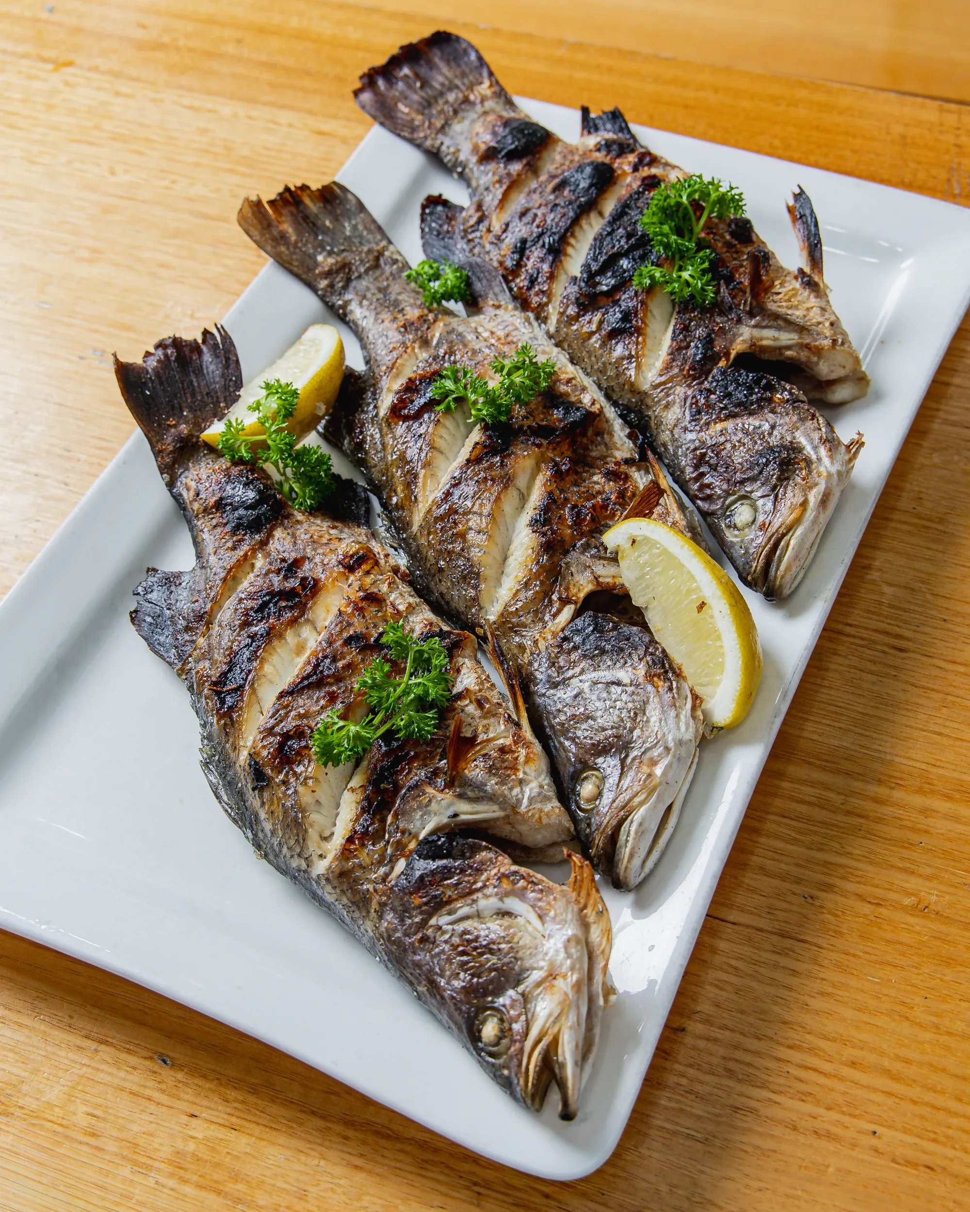 Grilled whole fish garnished with parsley and lemon wedges on a white rectangular plate, placed on a wooden table.
