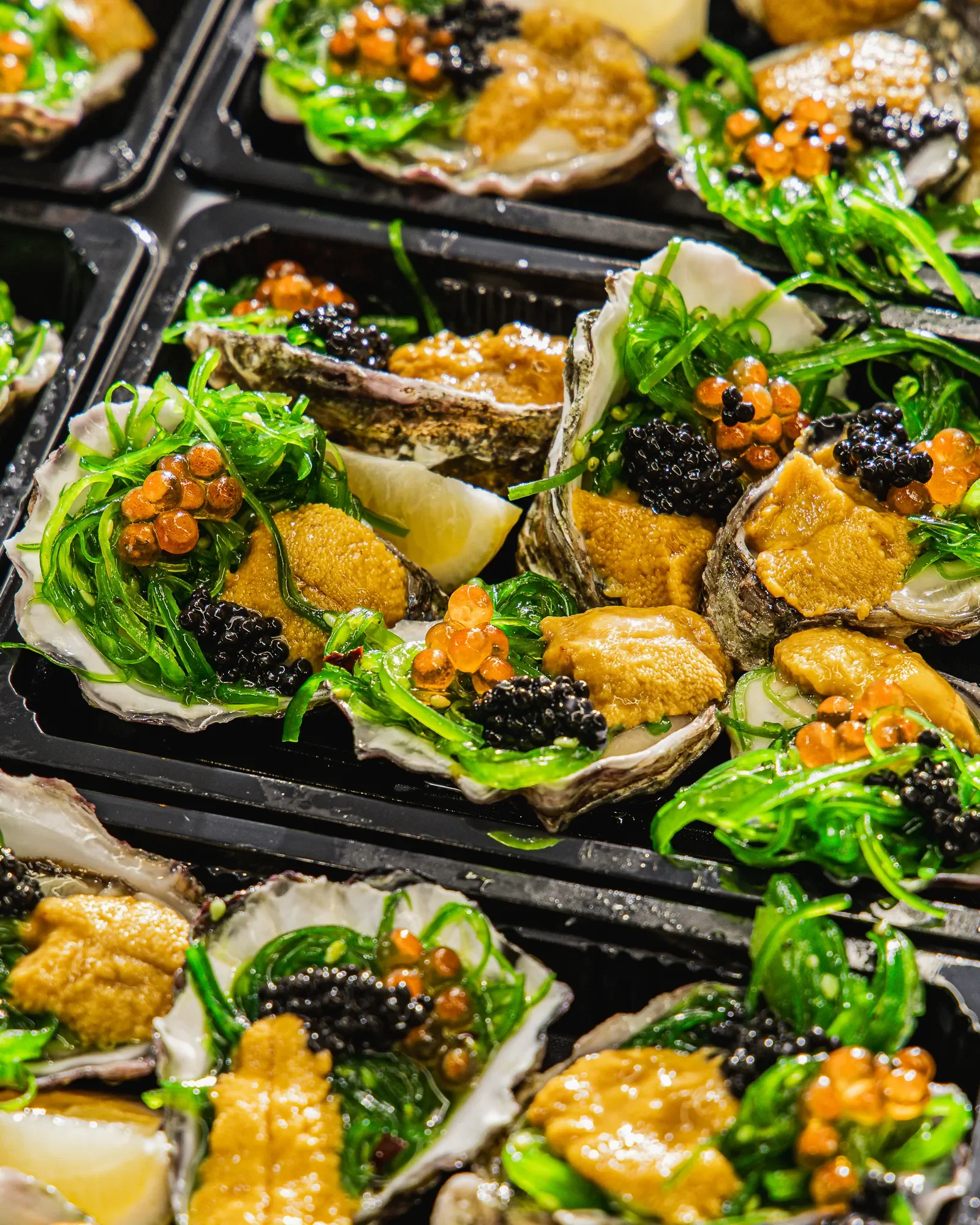 Close-up of oysters topped with seaweed, black caviar, salmon roe, and lemon wedge on a black tray.