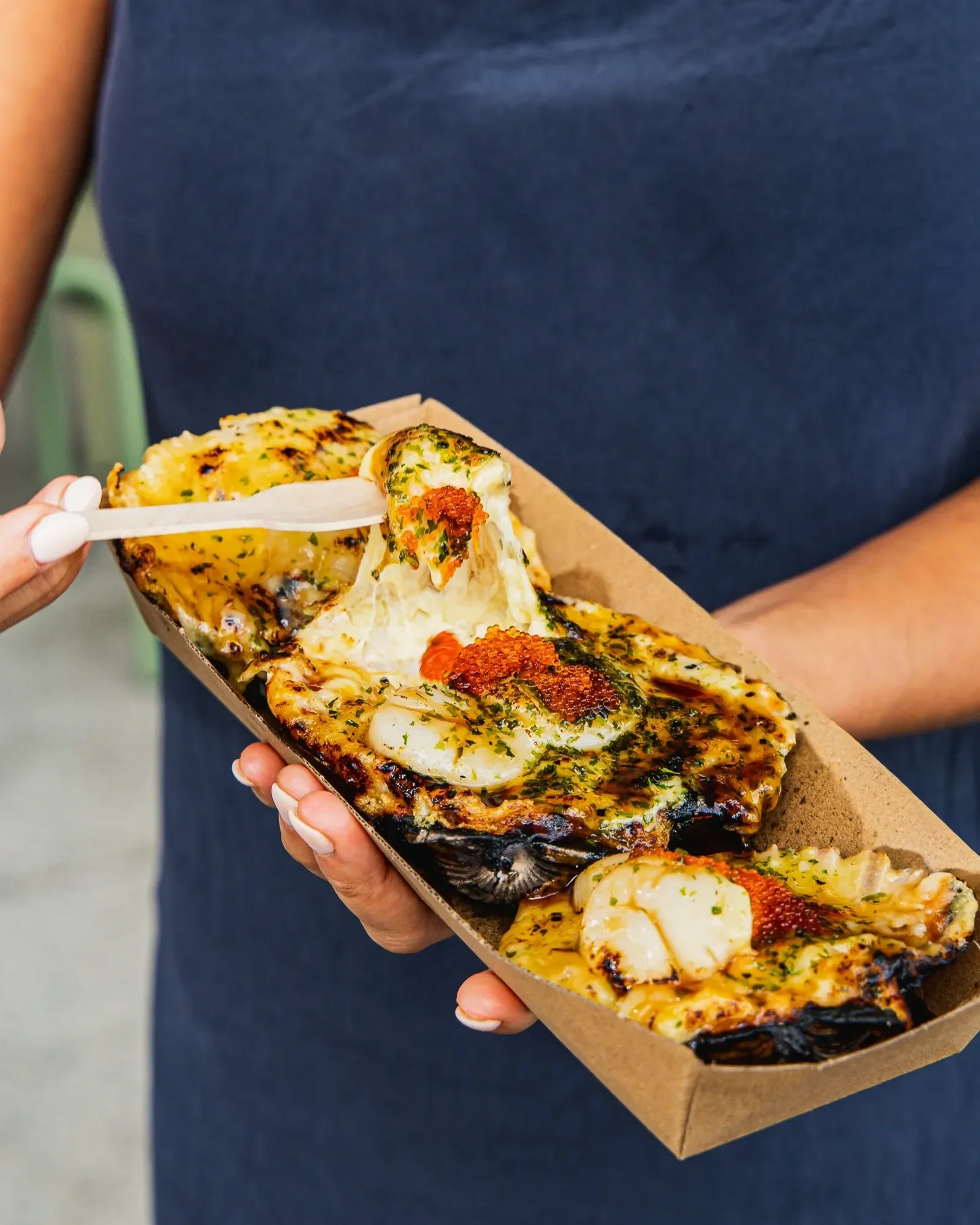 Close-up of a person holding a cardboard tray of seafood pizza with seafood toppings and herbs, using a white plastic fork.
