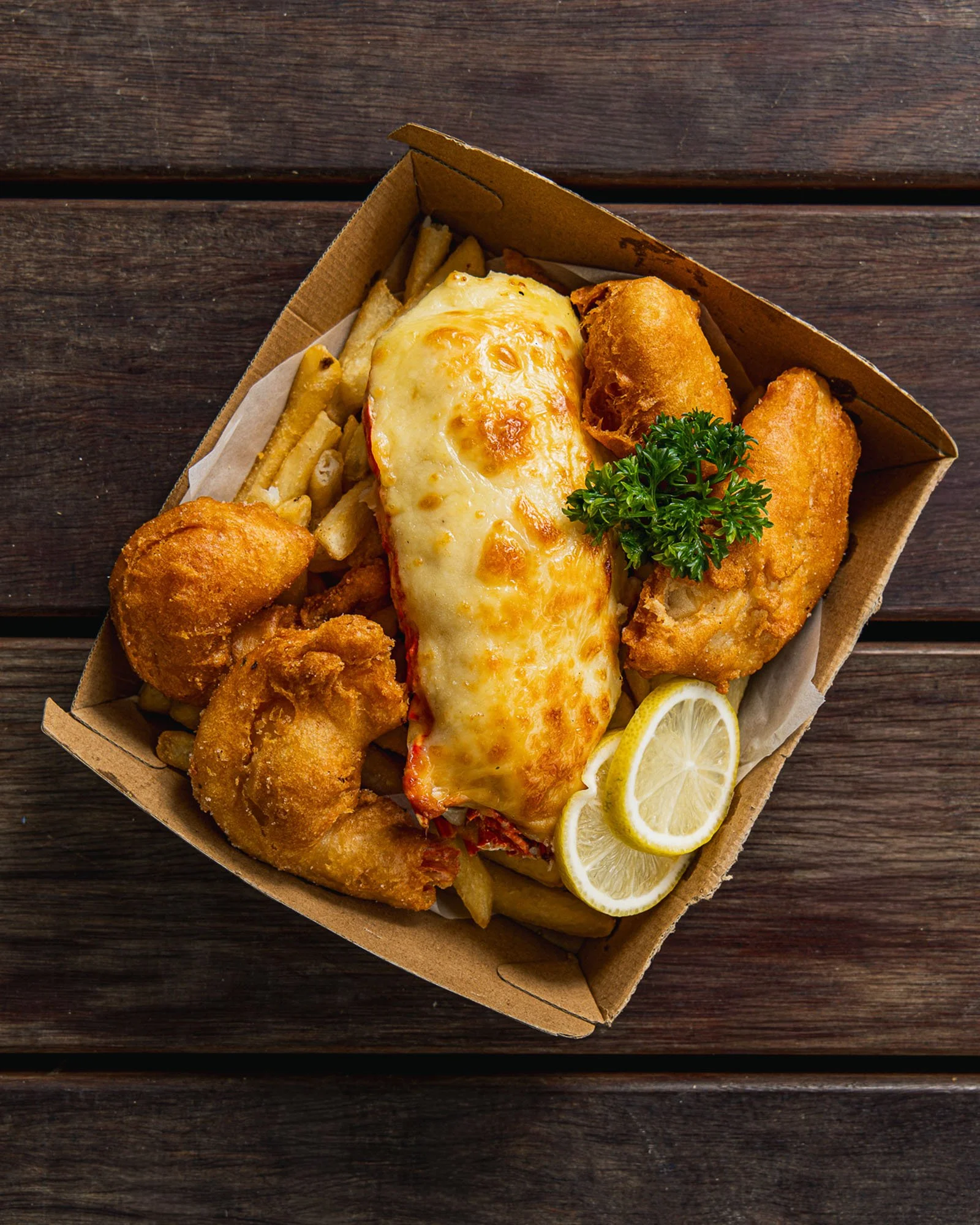 A takeout container filled with fried fish fillets, cheeseburger with melted cheese, French fries, lemon slices, and parsley on a dark wooden surface.