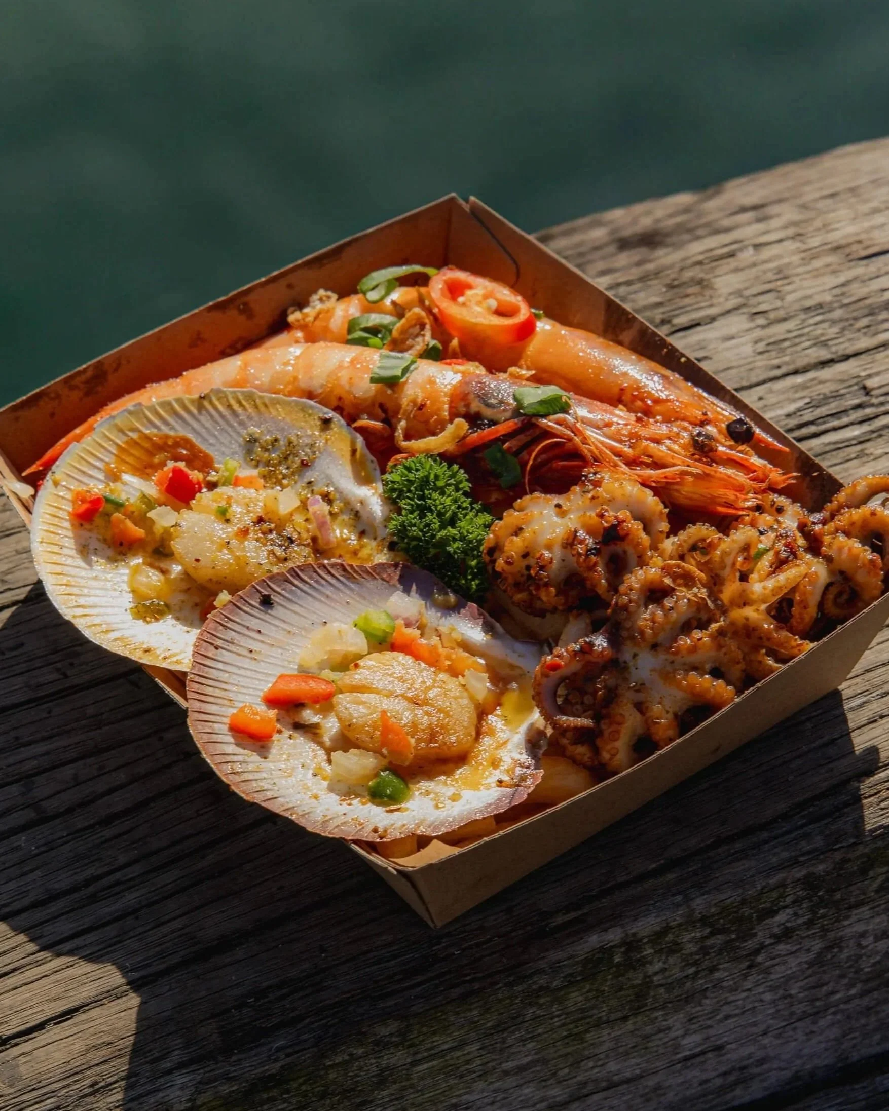 A seafood platter with grilled salmon, shrimp, octopus, stuffed crab, roasted potatoes, lemon wedges, and garnishes served on a rectangular tray on a marble surface.