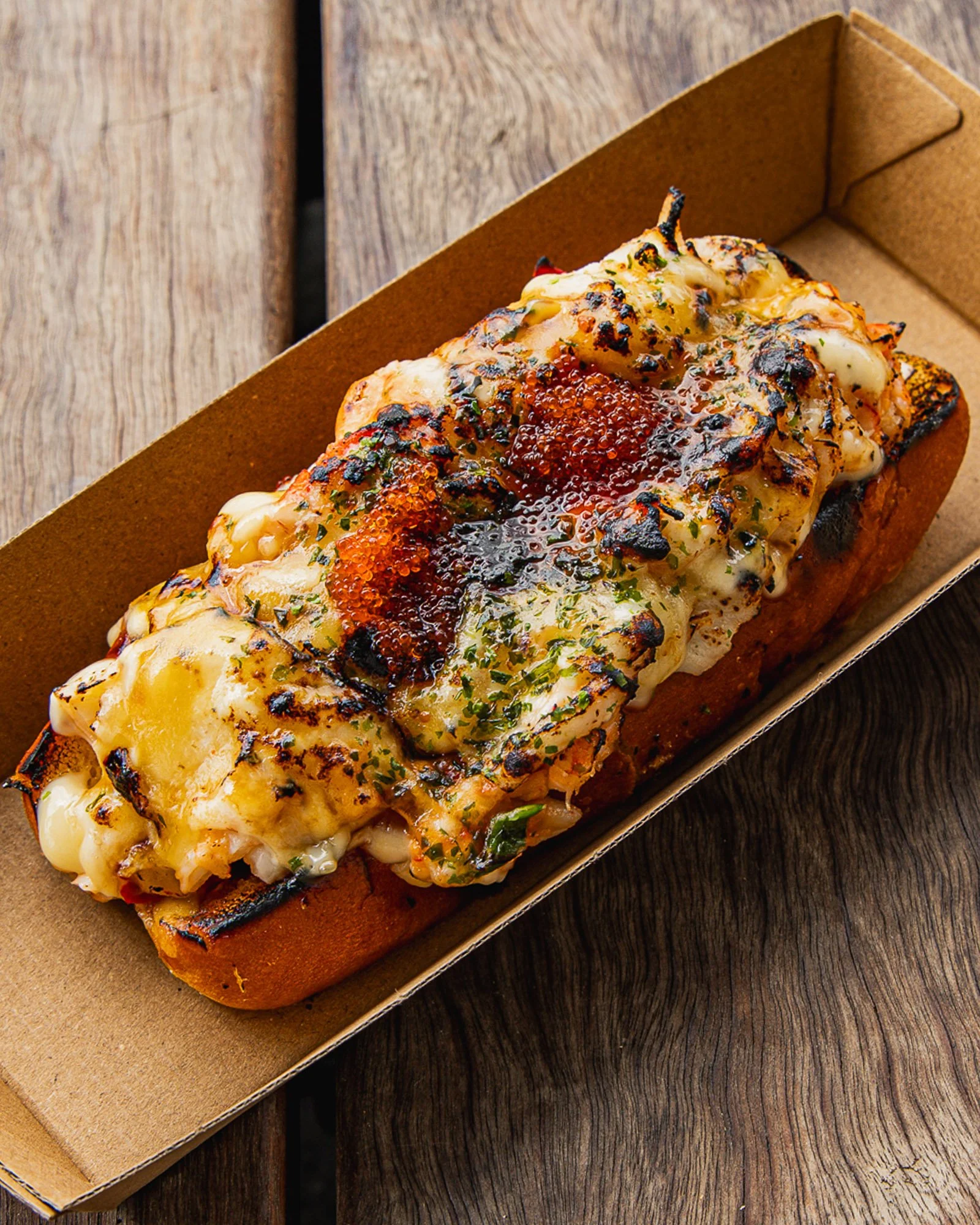 Close-up of a gourmet open-faced sandwich with melted cheese, salmon flakes, and herbs on toasted bread, served in a brown paper tray on a wooden surface.