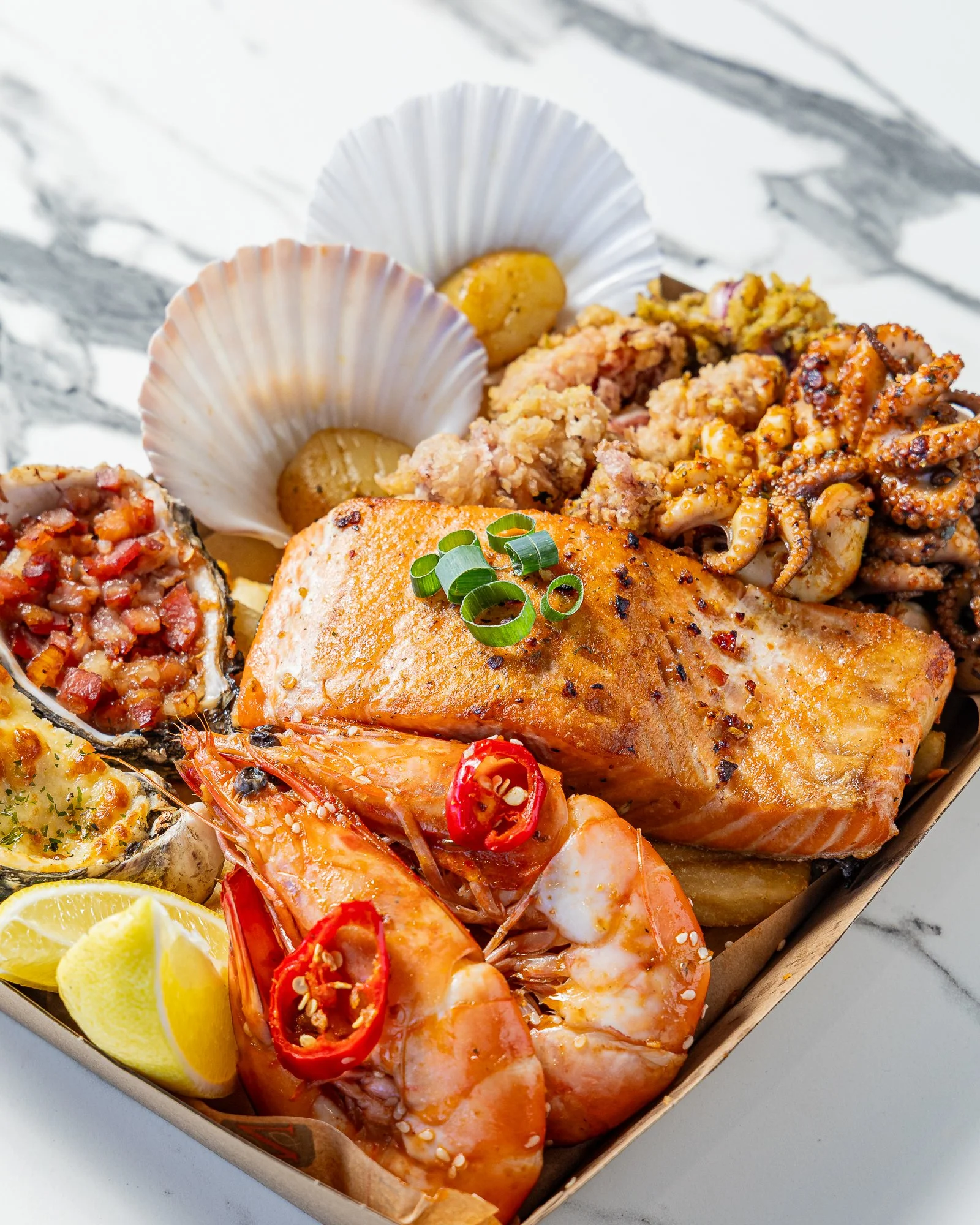 Seafood platter with grilled salmon, prawns, octopus, lemon wedges, and scallops, served on a tray.