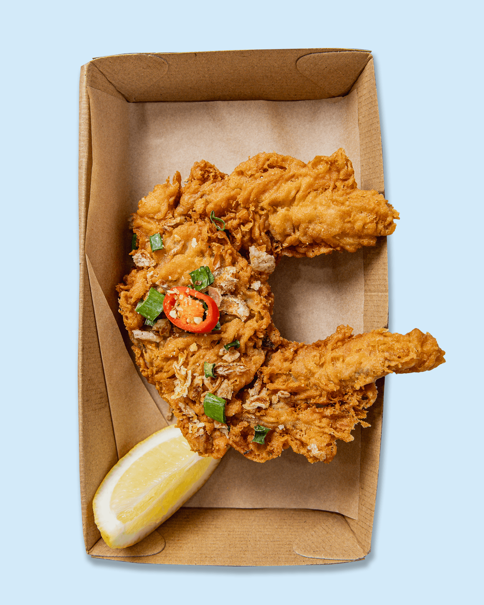 A cardboard takeout box containing fried chicken strips garnished with chopped green onions and sliced red chili, alongside a lemon wedge.