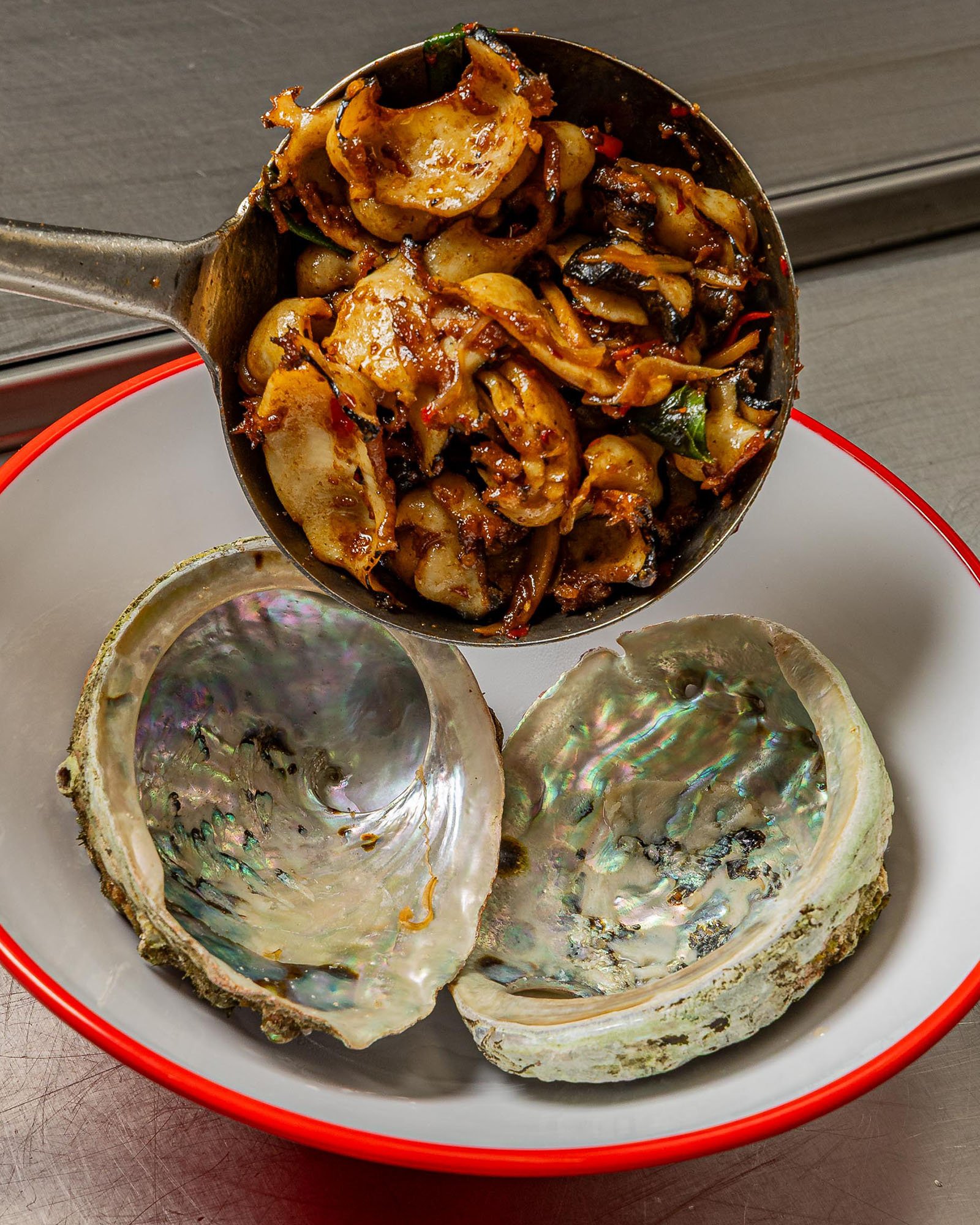 A blackened metal ladle filled with stir-fried vegetables, including onions and green peppers, above a bowl of empty oyster shells with iridescent interiors on a silver surface.