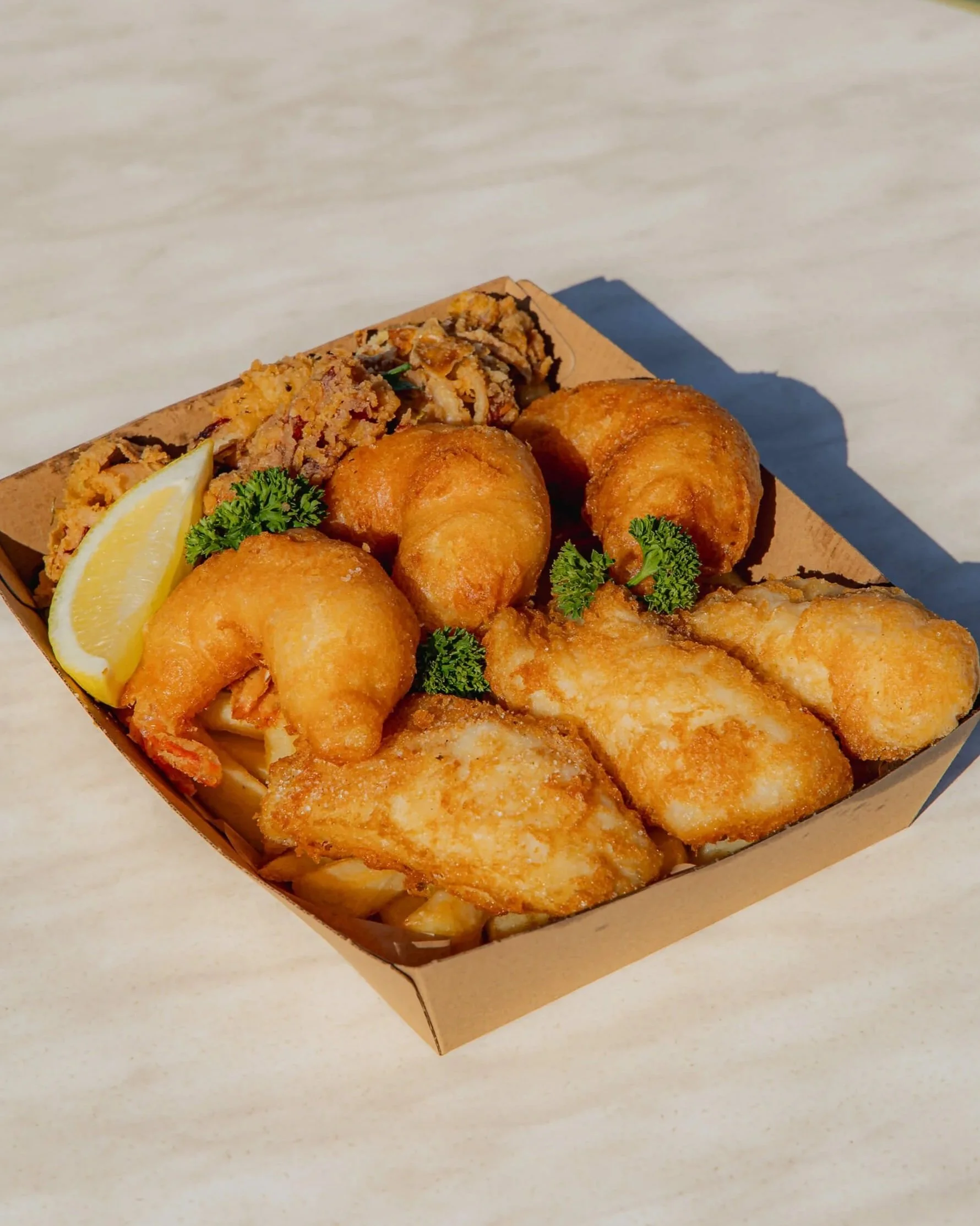 A basket of fried fish fillets garnished with lemon wedges and parsley, with a side of crispy breaded chicken wings, on a white marble surface.