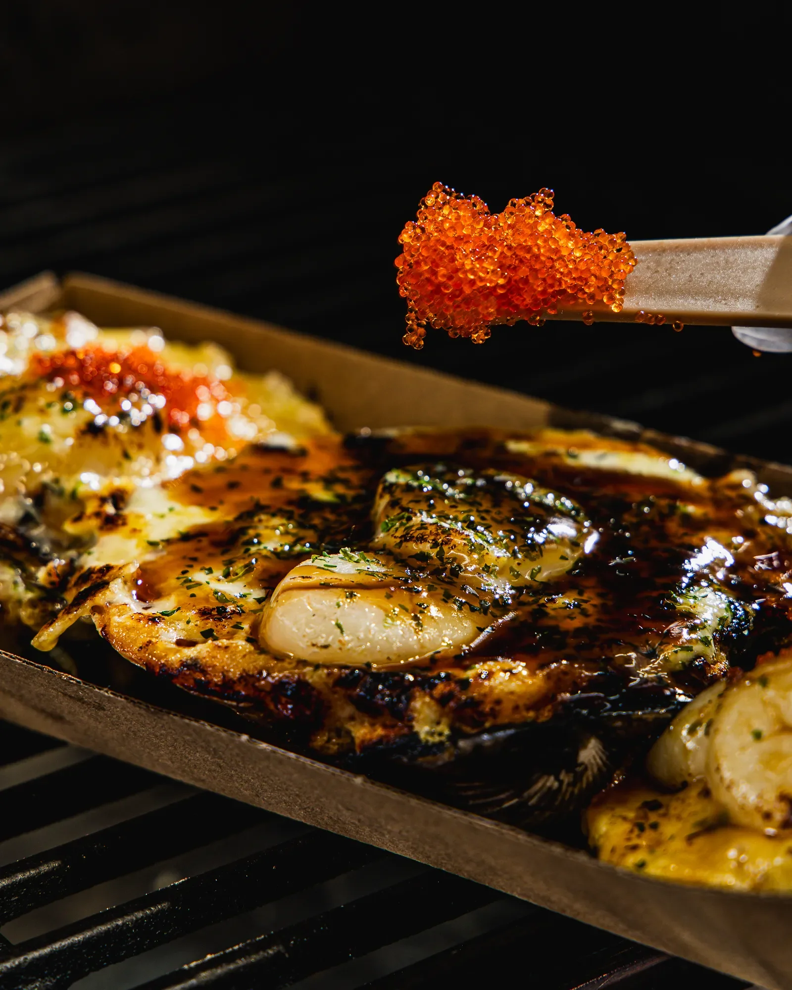 A close-up of fish being cooked on a grill with a piece of sushi with bright orange fish roe on a chopstick in the foreground.