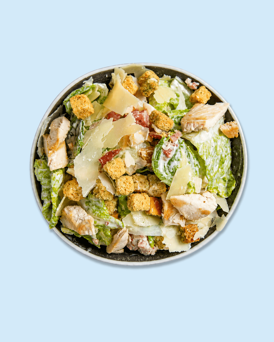 Top view of a Caesar salad in a bowl with lettuce, grilled chicken pieces, parmesan cheese, croutons, and bacon bits, on branded paper.